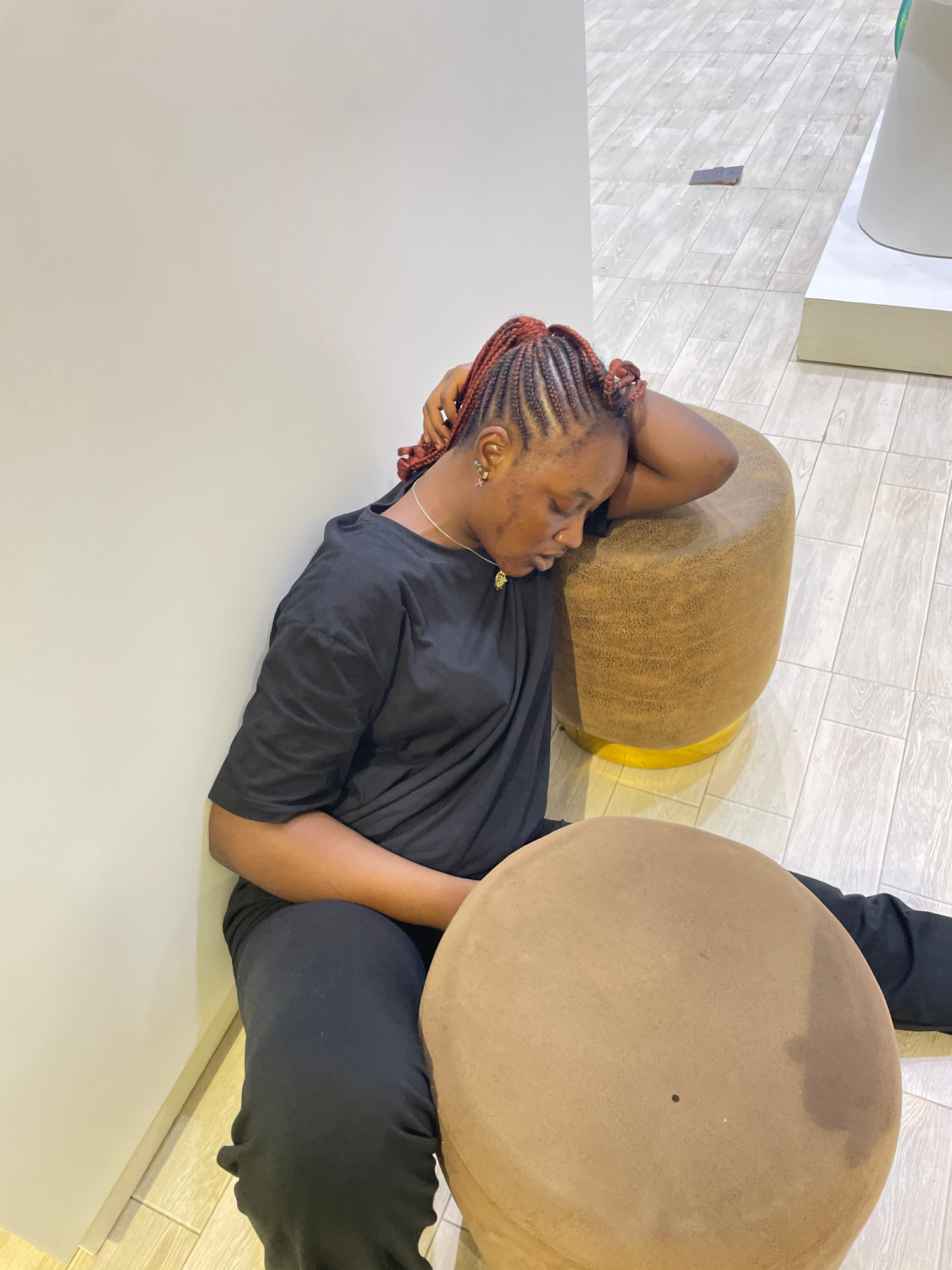 The image shows a person lying down on a light-colored surface, possibly a table or floor. The person is wearing a black t-shirt and black pants. Their arms are resting behind their head, and their eyes are closed, suggesting they are resting or sleeping. The person has braided hair with reddish-brown highlights and is wearing a silver necklace and earrings.
Behind the person are two round, ottoman-like objects. One is a textured, brown ottoman, and the other is a larger, flatter, light brown circular surface, possibly a coffee table or another ottoman. The background consists of a tiled floor with a pattern of light and dark grey rectangular tiles, and a partially visible wall.
The scene appears to be indoors, likely in a modern or minimalist setting given the clean lines and simple furniture. There are no strong indicators of the time of day or weather conditions. The overall mood conveyed is one of relaxation and repose. The location context provided is Unguwar Sarki, Nigeria.