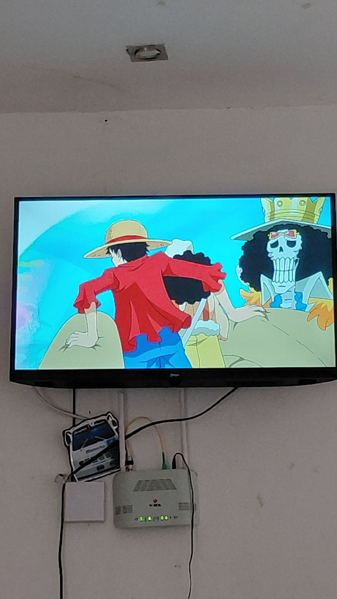 A flat-screen television displays an anime scene, featuring characters from "One Piece." On the wall below the television, a white Wi-Fi router is mounted, with wires connected to it. To the left of the router, a sticker of a white bus is affixed to the wall. The scene is illuminated by ambient light, and the overall environment appears to be a domestic setting in Santa Ana, Venezuela.