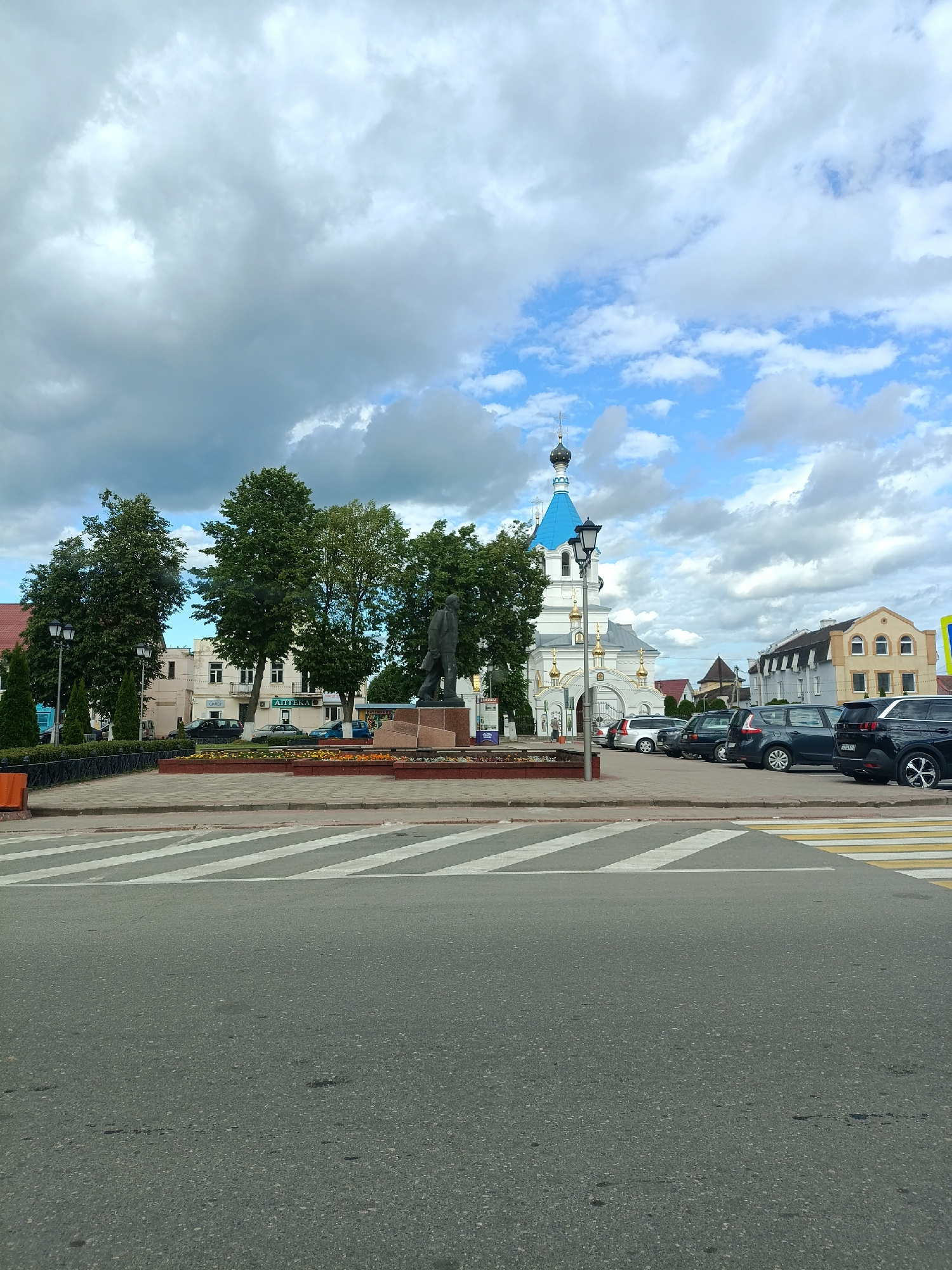 A low-angle, eye-level medium shot captures a public square with a prominent statue and a white church with a blue dome in the background. The foreground features a wide asphalt road with a pedestrian crosswalk marked by white stripes. To the left and right of the crosswalk, the sidewalk is visible, paved with light-colored stone. Beyond the sidewalk is a raised planter box filled with a mix of green foliage and small orange flowers. Behind the planter, a dark metal fence lines the perimeter of the square. A tall, dark metal lamppost stands on the left side of the planter.
In the mid-ground, a bronze statue of a standing man is positioned on a stone pedestal. Behind the statue, several mature trees with dense green foliage are scattered throughout the square, obscuring portions of the buildings. These buildings appear to be a mix of architectural styles, with some featuring light-colored facades and white window frames, while others are a darker color with arched windows.
The background is dominated by a white church with multiple golden domes and a prominent blue dome topped with a cross. The church entrance features ornate gold detailing. To the right of the church, a light yellow building with arched windows is visible. A row of parked cars, including silver, dark blue, and black SUVs and sedans, is situated along the side of the square, in front of the buildings. The sky above is a mix of bright blue patches and large, heavy grey clouds, suggesting potentially changeable weather. The overall scene depicts a public space in a European city, likely on a slightly overcast day.
