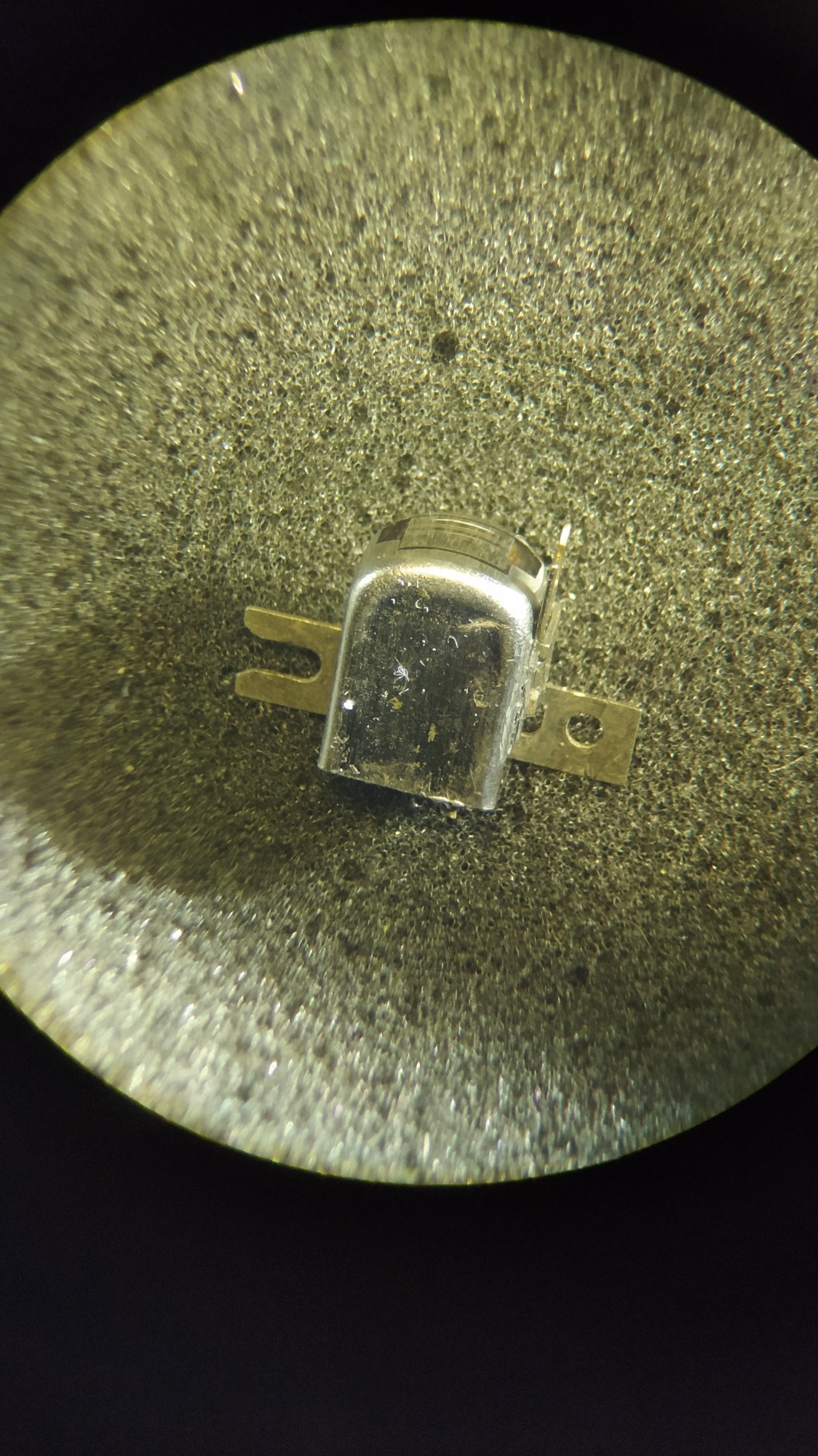 A close-up, microscopic view of a small, metallic electronic component is presented. The component is silver-colored and has a rectangular, rounded top housing. Two flat metal tabs extend horizontally from the sides of the housing, one on the left and one on the right. The left tab is U-shaped with a gap in the middle, while the right tab is a solid rectangular piece with a circular hole at its end. The component is positioned on a textured, gray surface, which appears to be a foam-like material with many small, irregular pores. The lighting in the image creates specular highlights on the metallic surface of the component and illuminates the porous texture of the background. The overall image is circular, with the top portion being brightly illuminated and the bottom portion fading into darkness. There is no information available about the location in Kyiv, Ukraine, or any context regarding people, actions, or interactions.