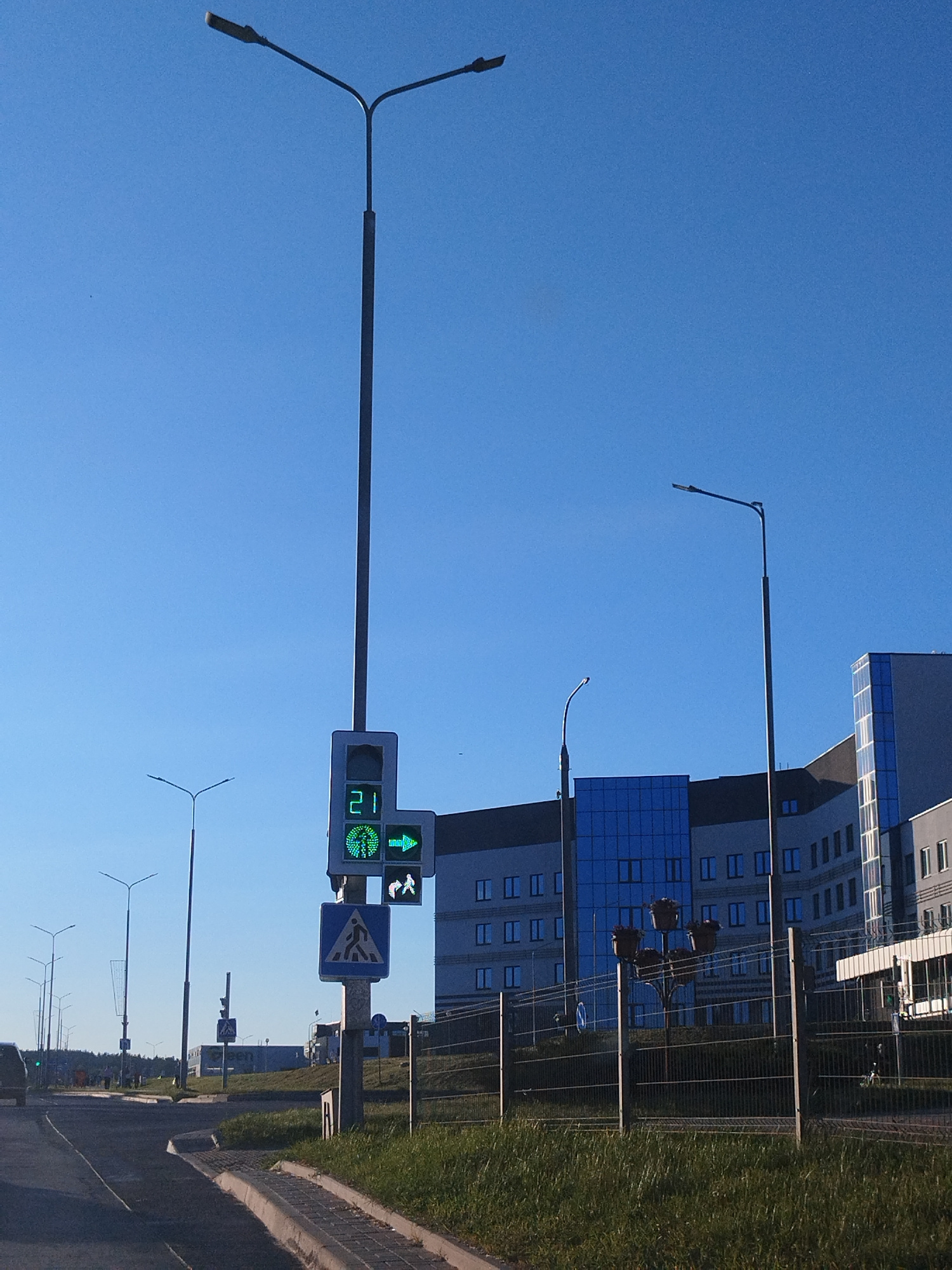 A traffic light with a countdown timer showing 21 seconds is illuminated in green, indicating a go signal for traffic. Next to it, a pedestrian crossing signal displays a green walking figure and a green right-pointing arrow. Below the traffic lights, a blue rectangular sign with a white pedestrian symbol is mounted on a pole. The scene is set on a sunny day with a clear blue sky. In the background, a modern building with blue-tinted windows and a dark facade is visible. A metal fence with wire mesh runs along the foreground, separating a grassy area from a paved road. Several streetlights are present throughout the scene.