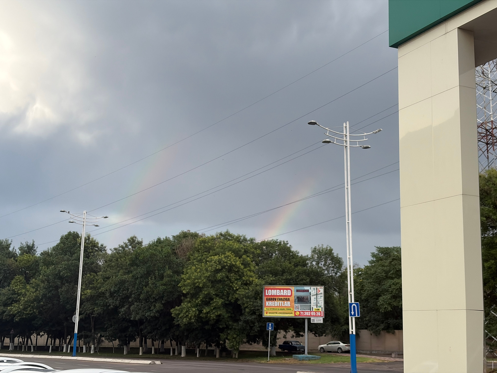 This image captures a cloudy sky with two visible rainbows arching over a row of lush green trees. In the foreground, a modern building with a cream-colored facade and a green roof extends into the frame on the right. A white street lamp with multiple lights stands prominently in the middle ground, accompanied by power lines stretching across the sky.
A billboard is visible amongst the trees, featuring a bright advertisement for "LOMBARD" which appears to offer loans against valuables and technical equipment, with a phone number "71 202 05 26" and address "SHASTRI CHORRAHASI" printed on it. Two cars are parked behind the billboard, one dark blue and one silver. In the lower part of the image, the tops of parked white cars are visible, suggesting a parking lot or roadside setting. A blue and white traffic sign with a U-turn symbol is placed near the street lamp.
The presence of rainbows and the overcast sky suggest recent rain or ongoing precipitation, likely occurring during daylight hours. The setting appears to be an urban or suburban area in Tashkent, Uzbekistan, given the context provided. The overall mood is serene despite the cloudy weather, with the vibrant rainbows adding a touch of natural beauty to the scene.