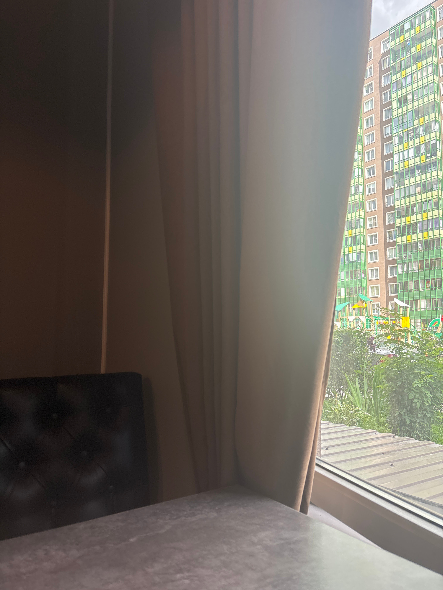 The image captures a view from a window, looking outwards and slightly downwards. In the background, a tall, multi-story building dominates the scene. Its facade is a mosaic of light brown panels and a grid of windows with light green frames, some of which contain yellow accents. The building appears to be an apartment complex or office building. To the right of the building, lush green trees and foliage are visible, suggesting an outdoor environment. The sky above is overcast, indicating a cloudy day.
In the foreground, a thick, light brown curtain is draped, partially obscuring the view. The curtain has a soft texture and is gathered, creating folds and shadows. To the right, a section of what appears to be a textured wall or furniture, possibly upholstered, is visible, with a rough, light grey surface. At the bottom right of the image, a dark, tufted piece of furniture, likely a chair or headboard, is partially visible. Its surface is dark and has a button-tufted pattern.
The overall impression is an indoor scene looking out at an urban landscape with greenery. The lighting suggests it is daytime, possibly late morning or afternoon, given the diffused light from the cloudy sky. There is no visible text in the image. The setting appears to be residential, given the view of an apartment building and the interior elements of curtains and furniture.