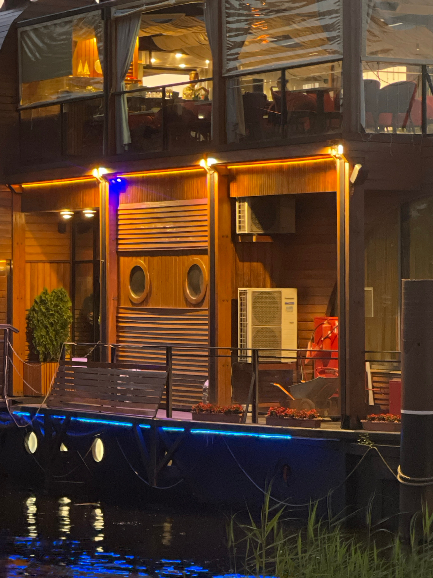The image shows a multi-level wooden structure resembling a houseboat or a floating restaurant docked on a body of water, possibly a canal or river in Saint Petersburg, Russia. The structure is illuminated by warm orange and cool blue LED lights. The upper levels have large windows with sheer white curtains, revealing glimpses of a dining area with tables and chairs. The middle level features wooden paneling, two circular porthole windows, and what appears to be a large sliding door or shuttered section. An outdoor air conditioning unit is mounted on the wall of this level. Below, a dark-colored deck has a wooden bench and railings. The water reflects the lights from the structure and some aquatic plants are visible along the edge. The overall impression is that of a tranquil evening scene at a waterfront establishment.
