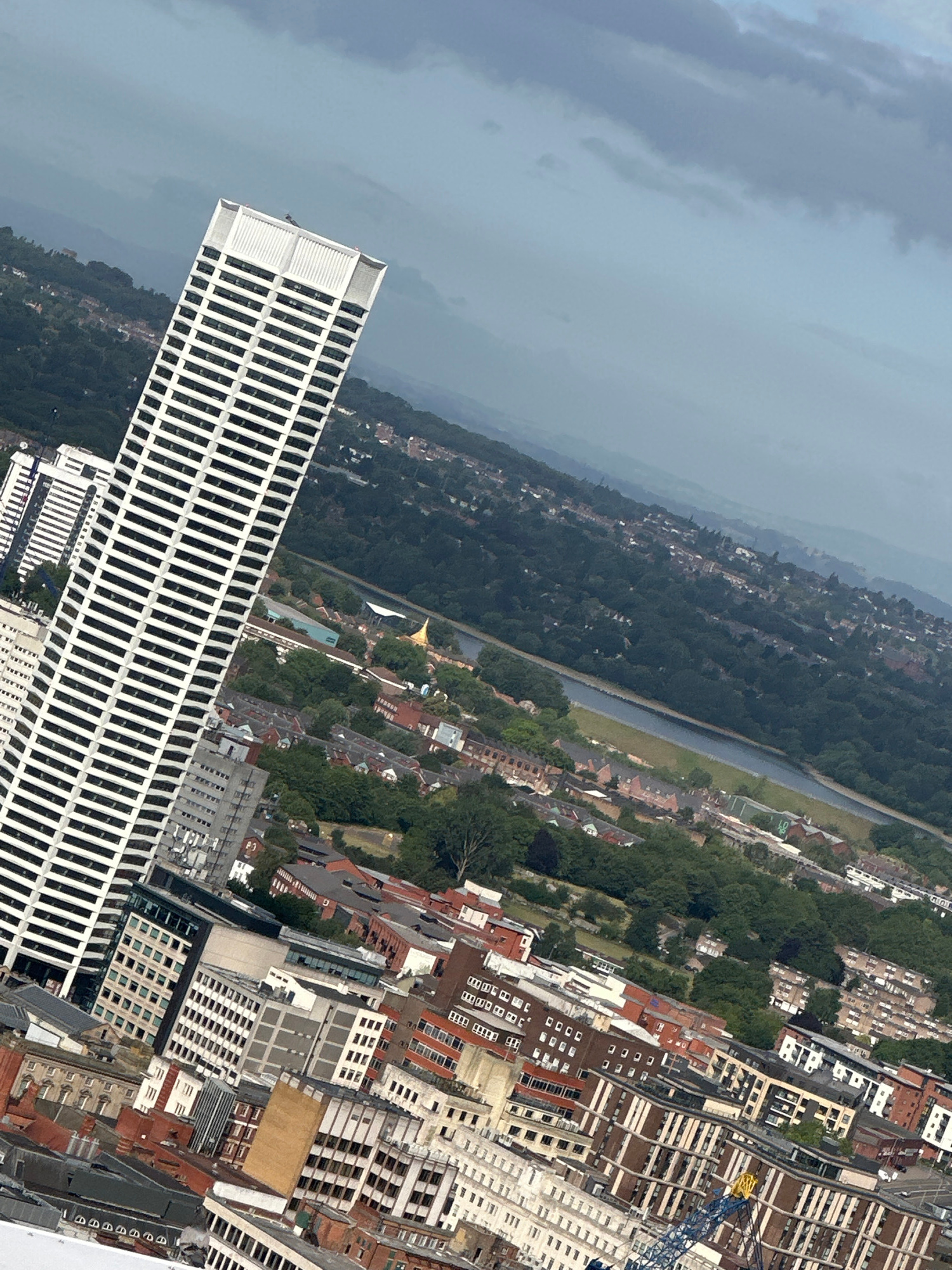 This is an aerial, high-angle, tilted view of a cityscape with a focus on urban architecture and natural elements. A prominent white, rectangular skyscraper with many vertical window panes dominates the foreground on the left. To the right of this skyscraper, the image shows a dense collection of various buildings, including apartment blocks and commercial structures, with a mix of red-tiled roofs and lighter facades. A bright yellow construction crane is visible amidst these buildings.
Further into the background, a body of water, likely a river or canal, winds through the scene, bordered by a green embankment. Beyond the water, a vast expanse of trees indicates a significant green area within or adjacent to the city. In the far distance, rolling hills are shrouded in a grey, hazy atmosphere, suggesting overcast or stormy weather. The sky itself is filled with heavy, dark clouds, contributing to a subdued and somewhat dramatic mood.
The time of day appears to be daytime, given the illumination of the buildings and landscape, but the heavy cloud cover prevents direct sunlight and casts a muted light. There are no visible people in the image. The scene captures a juxtaposition of dense urban development with natural green spaces and a potentially approaching weather front, creating a layered and complex urban landscape. The location context identifies the city as Birmingham, United Kingdom.