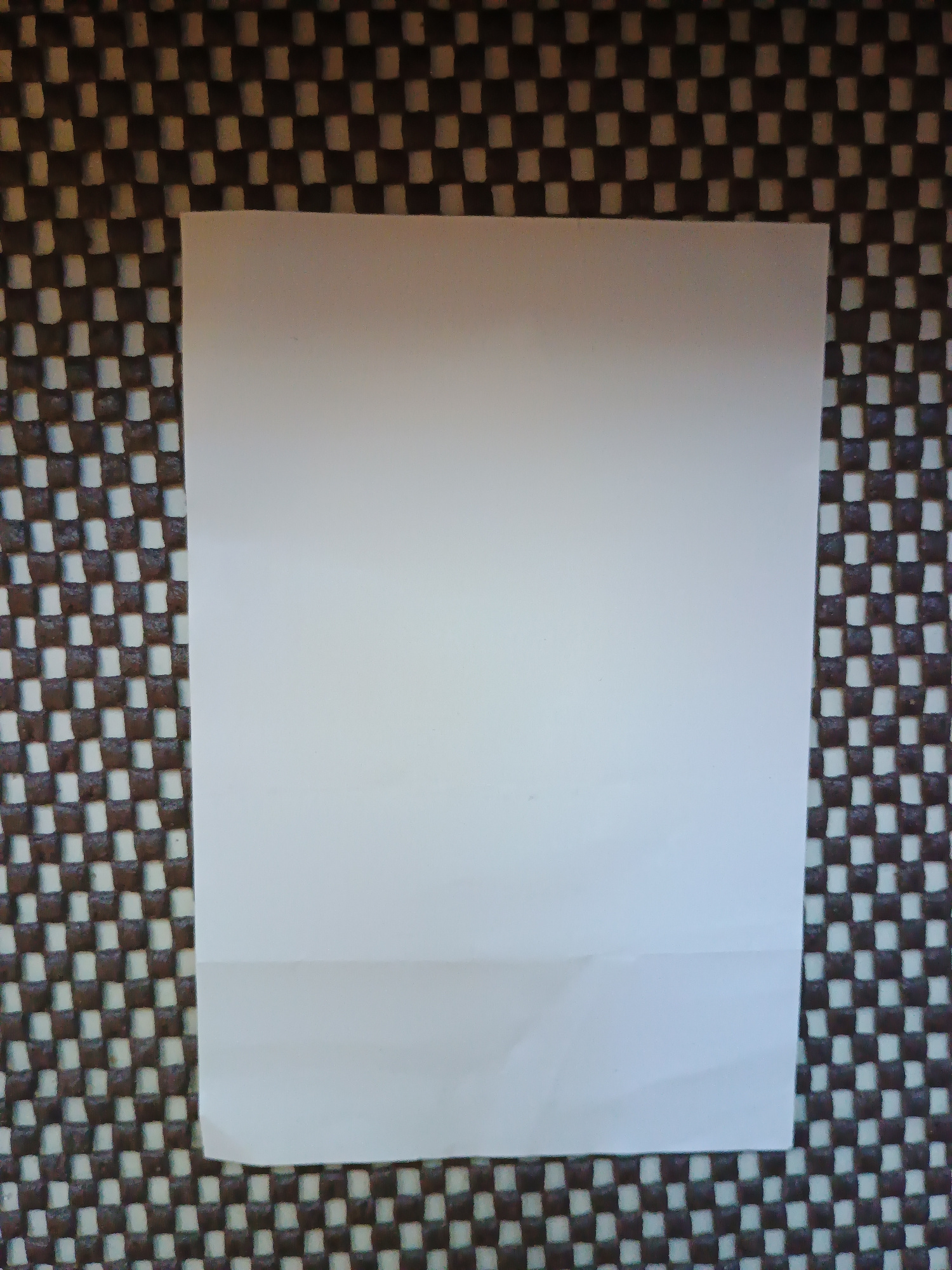 A white sheet of paper is placed on a patterned surface. The surface appears to be a woven material with a repeating checkerboard pattern of dark brown and light beige squares. The paper is positioned centrally and takes up a significant portion of the frame. The bottom edge of the paper has a slight crease or fold. No people or actions are depicted. The scene is indoors, with neutral lighting conditions. The location in Zaporizhzhia, Ukraine, cannot be determined from the visual information provided.