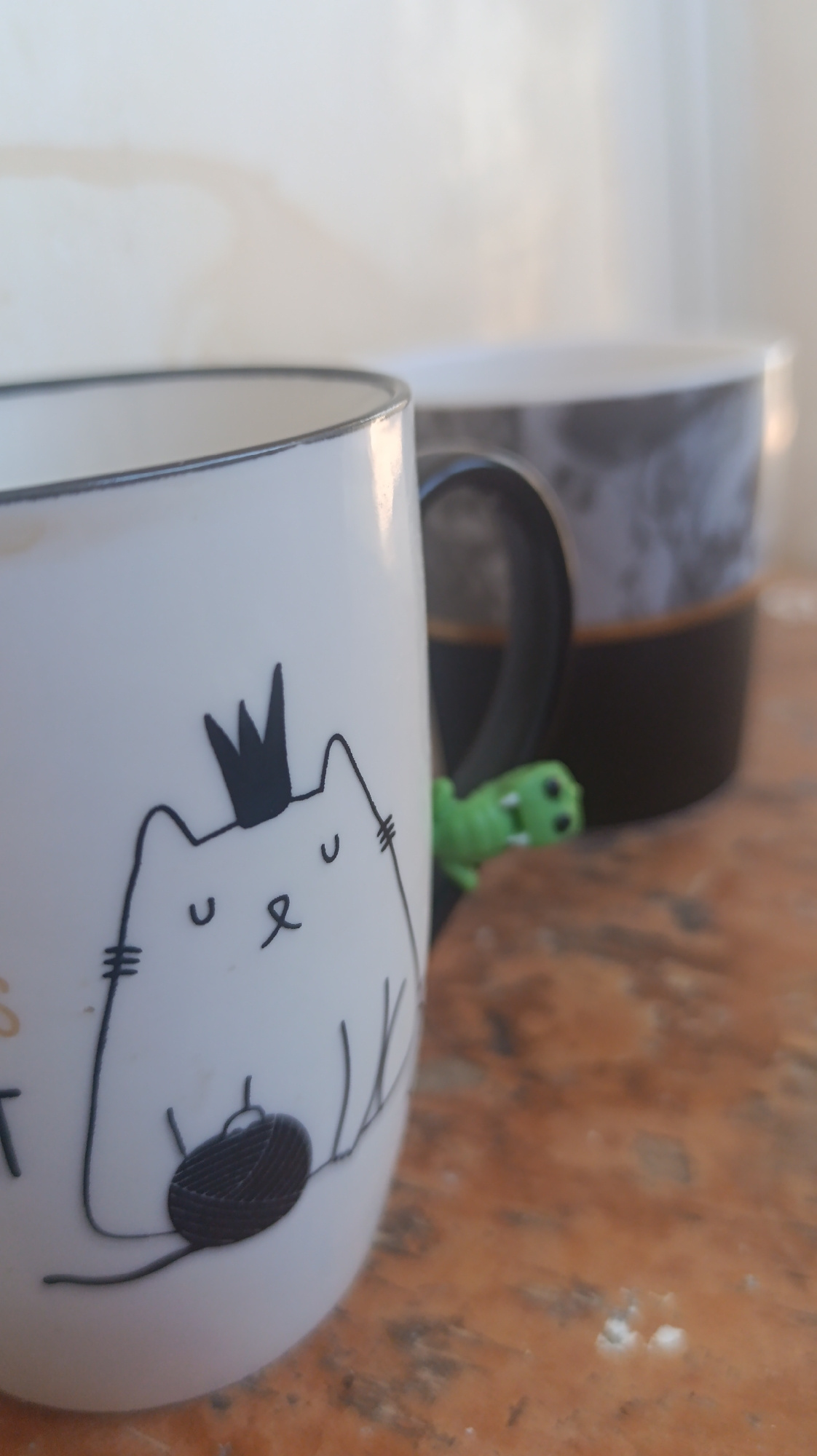 The image features two mugs on a wooden surface. The mug in the foreground is white with a black rim and a black handle. It has a black line drawing of a cat wearing a crown and holding a ball of yarn. The cat has simple eyes and a nose. To the right of this mug, a small green plastic dinosaur figurine is perched on the handle.
Behind the white mug is another mug, partially visible. It has a black exterior with a marbled gray pattern and a gold band near its rim.
The background is out of focus, showing a white or light-colored wall with some faint smudges. The lighting suggests it might be daytime, possibly morning or afternoon, with natural light illuminating the scene from the side. The overall impression is a casual, indoor setting, perhaps on a table or shelf. There is no discernible text in the image, aside from a partial word in gold on the left side of the foreground mug which is obscured. The focus is primarily on the mugs and the charming cat illustration.