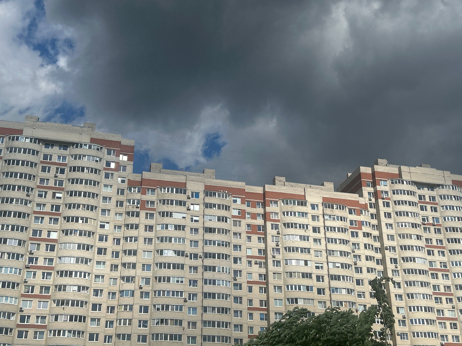 This media file depicts a large, multi-story apartment building under a dramatic, overcast sky. The building is constructed with light-colored concrete panels and features decorative red brick accents around the upper floors and window frames. The architecture suggests a residential block, typical of urban development. The windows are numerous and uniformly arranged, with many featuring balconies or curved bay windows, indicating individual apartment units.
The sky is dominated by dark, heavy storm clouds, suggesting an approaching or ongoing rain shower. Patches of blue sky are visible in the upper left quadrant, hinting at a dynamic weather system. The overall mood is somber and perhaps a bit foreboding due to the dark clouds. The time of day is difficult to ascertain precisely but appears to be daytime, given the illumination on the building and the visible sky.
The setting is likely a residential area in Saint Petersburg, Russia, as indicated by the architectural style and the context provided. There are no people clearly visible, but the presence of lights in some windows suggests occupancy. Some trees with lush green foliage are visible in the lower right corner, contrasting with the grayness of the sky and the uniformity of the building. Electrical wires are faintly visible strung across the upper portion of the building. No visible text is present in the image.