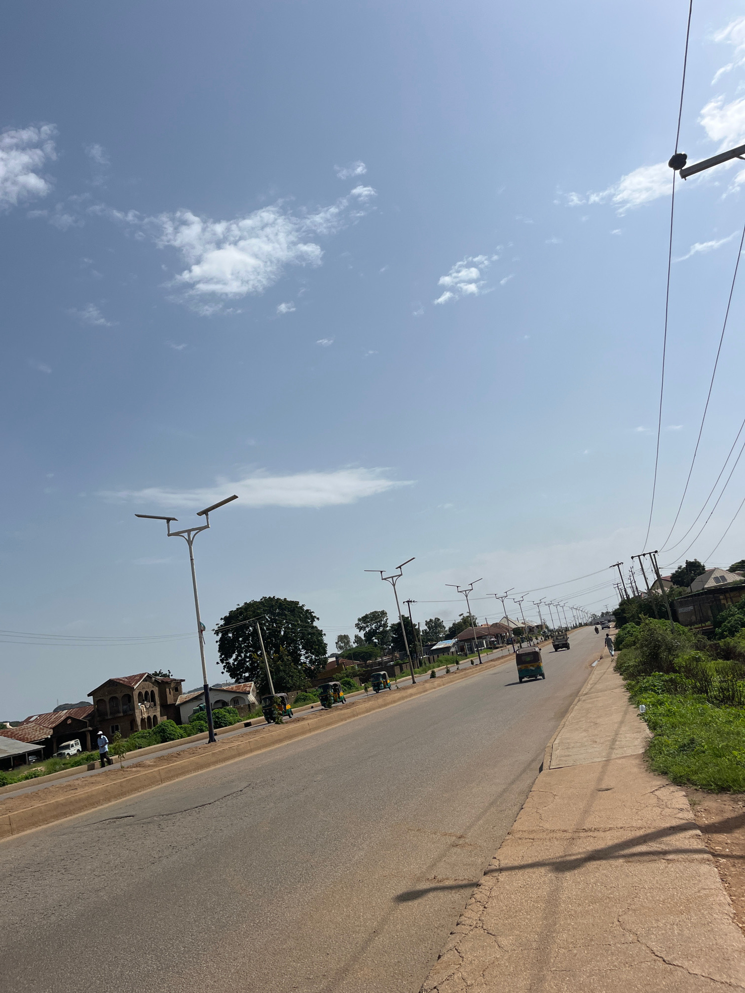 This is a ground-level, eye-level, medium shot capturing a street scene in Bukuru, Nigeria. The paved road, appearing to be a main thoroughfare, stretches into the distance, curving slightly to the right. The road surface exhibits some wear and tear, with visible cracks and patches. On the left side of the road, a sidewalk or paved path runs alongside, also showing signs of age and cracking. Beyond the sidewalk, there are buildings with corrugated metal roofs, typical of residential and commercial structures in the area. A large tree with dense foliage stands prominently in the mid-ground on the left.
Several vehicles are present on the road, including at least two auto-rickshaws, one white SUV, and other cars and trucks in the distance. A lone figure, possibly a pedestrian, is walking on the sidewalk on the right side of the road. The environment is characterized by a clear blue sky with scattered white clouds. Numerous streetlights with a modern, dual-headed design are positioned along both sides of the road, indicating infrastructure development. Electrical power lines are visible, strung between poles, with one set running diagonally across the upper right portion of the frame. The general ambiance suggests a sunny day in a developing urban or semi-urban area.