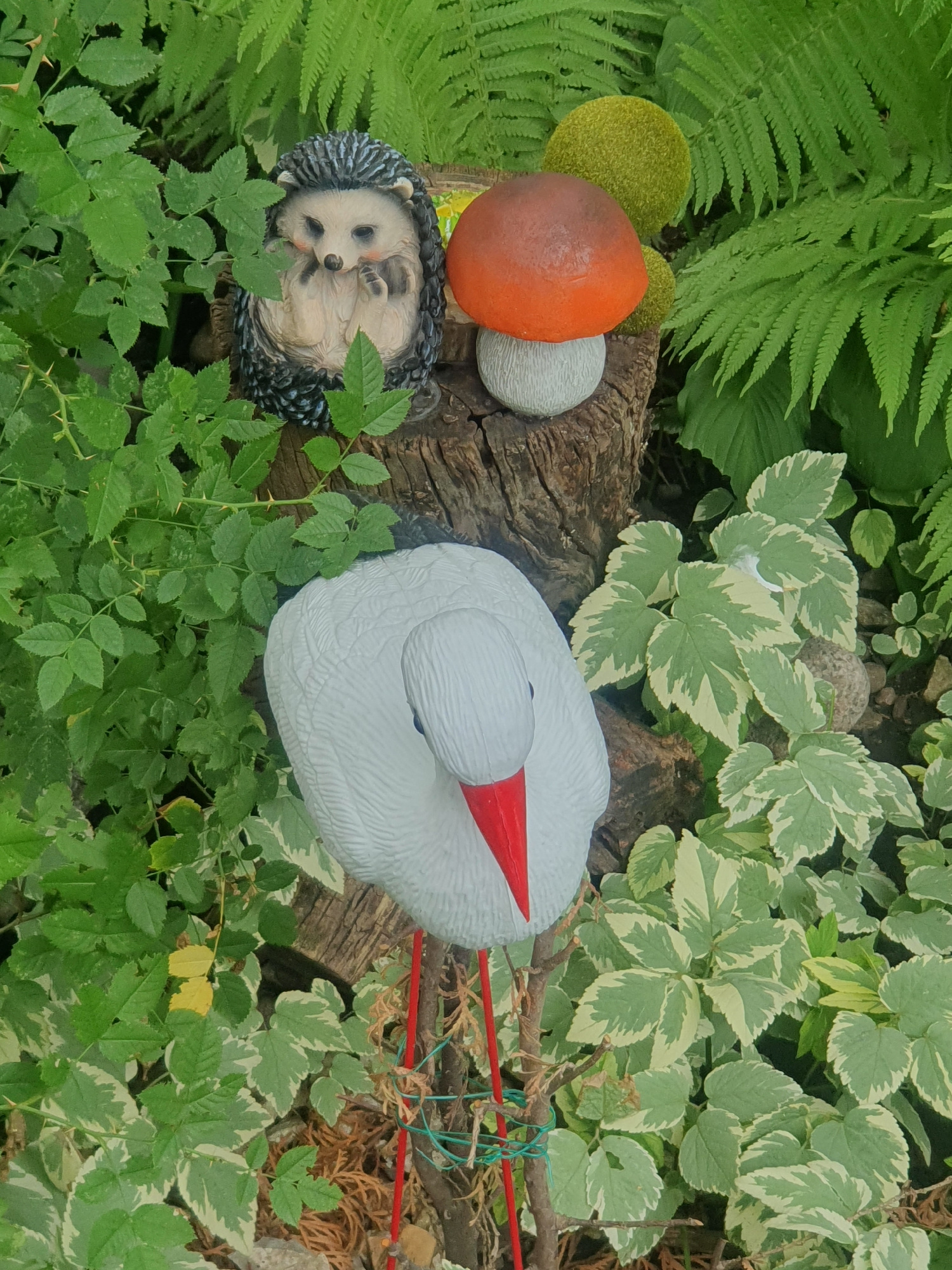 This image depicts a garden scene with several decorative elements. In the upper left, a ceramic hedgehog figurine is partially obscured by lush green foliage. To its right, a decorative mushroom figurine with an orange cap and a light gray stem stands on a weathered tree stump. Behind these, a vibrant green fern frond is visible. In the foreground, a white ceramic stork figurine with a red beak and red legs is the central subject. The stork is positioned among a variety of green and variegated green and white leaves. The overall setting appears to be a garden or a naturalistic outdoor space, possibly a park or residential garden. The specific location in Almaty, Kazakhstan is not discernible from the visual information provided.