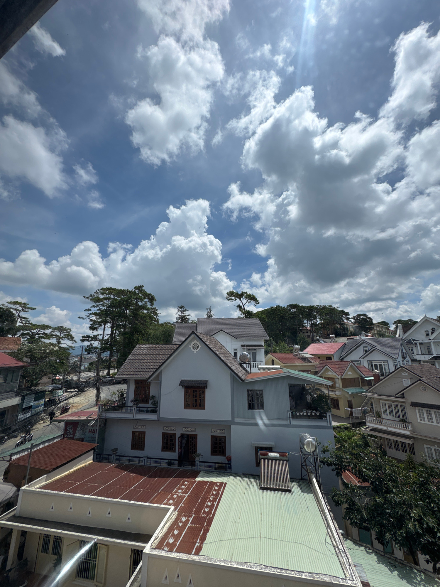 This image shows a view from a high vantage point, looking out over a residential area with a hilly landscape. The sky is filled with dramatic, cumulus clouds, with a bright sun casting lens flare in the upper right portion of the frame. The architecture suggests a temperate climate with pitched roofs and balconies. The buildings are of various sizes and colors, predominantly white, grey, and yellow, with many featuring terracotta tile roofs.
In the foreground, several rooftops are visible. The closest rooftop is a light-colored, flat structure with a green corrugated metal roof. To the left of this, another rooftop is partially visible, with a rusty, corrugated metal surface. Further back, a larger building with a light blue facade and white trim is prominent, featuring multiple windows, balconies, and a solar water heater on its roof.
Behind this building, a street is visible, lined with more houses and trees. The trees are tall and appear to be pines, characteristic of the Da Lat region. In the distance, the landscape continues with more buildings and greenery, suggesting a densely populated but naturally rich environment. The overall impression is of a sunny day in a picturesque, elevated urban setting. The presence of a solar water heater is common in many Vietnamese households, and the architectural style is consistent with residences found in cities like Da Lat, known for its French colonial influence and cooler climate.