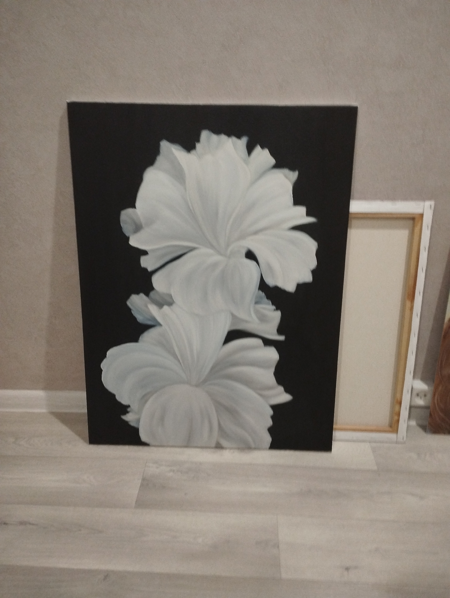 The image displays a vertical painting featuring two large, white, abstract flowers against a black background. The flowers are rendered with soft, rounded petals and a sense of depth. The painting is positioned on a light-colored wooden floor, leaning against a textured beige wall. To the right of the main painting, another canvas is visible, framed in white wood with a beige surface. The overall scene appears to be an indoor setting, possibly a room in a home or studio. No people are present in the image. The location of Balakovo, Russia is not discernible from the visual information provided in the image.