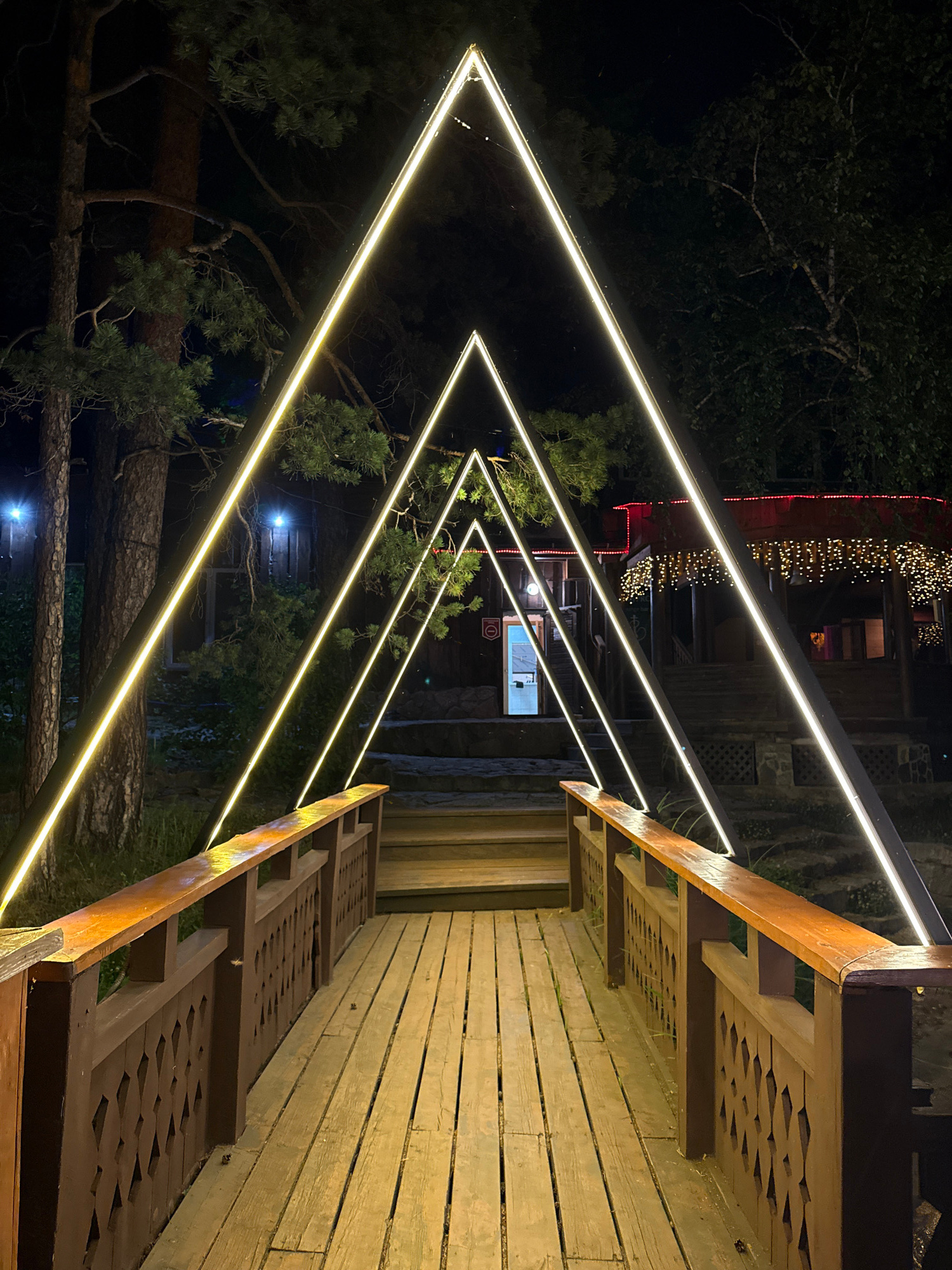 The image depicts a wooden bridge at night, illuminated by several triangular geometric light installations. The bridge has a wooden deck and a dark brown wooden railing with a patterned design. In the background, there are trees and a wooden building with festive string lights. The location is identified as Zolotoi Plyazh, Russia. The scene is serene and appears to be an outdoor recreational area. There are no people or animals visible in the image.