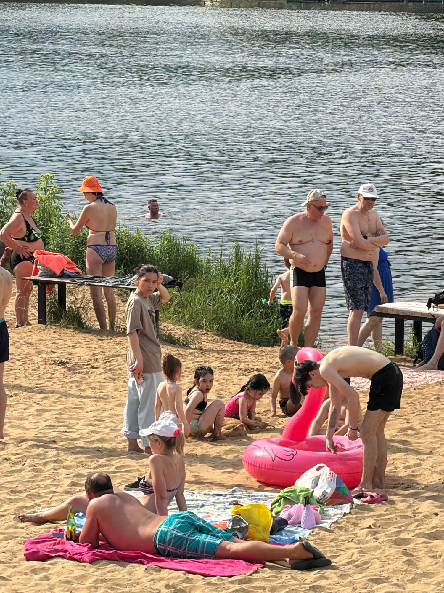 The image depicts a sunny day at a sandy beach on a lake in Malakhovka, Russia. Several individuals, including adults and children, are present. Some are in the water, while others are on the sand.
In the foreground, a man is lying on his back on a pink towel, facing away from the camera. He is wearing checkered green and blue shorts and has a bottle of beer next to him. A woman in a striped bikini is seated next to him.
Further back on the sand, a group of children are playing. A large, inflatable pink flamingo float is in the center of the image. Several towels and beach bags are scattered around.
In the middle ground, two women in swimwear are standing near a wooden table. One is wearing a green and black patterned bikini and the other an orange hat and blue bikini. Another woman in a tan t-shirt and light-colored pants is standing on the sand, looking towards the camera.
In the background, several men are standing near the water's edge, some with their arms crossed. A man is swimming in the lake. The lake is calm with ripples on the surface. Green foliage lines the edge of the water. The overall scene suggests a casual day of recreation.