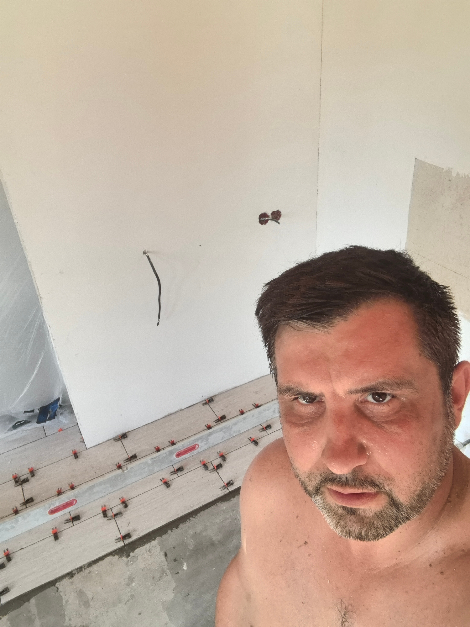 A man is taking a selfie in what appears to be a room undergoing renovation. The man, who is shirtless, has dark hair and a beard and is sweating. He is looking directly at the camera with a serious expression. In the background, the walls are painted white, and there are exposed electrical outlets and a hanging wire. The floor is partially covered with light-colored tiles, many of which are held in place by red and black leveling clips. The floor also shows areas of exposed concrete. The scene suggests construction or DIY work is in progress.