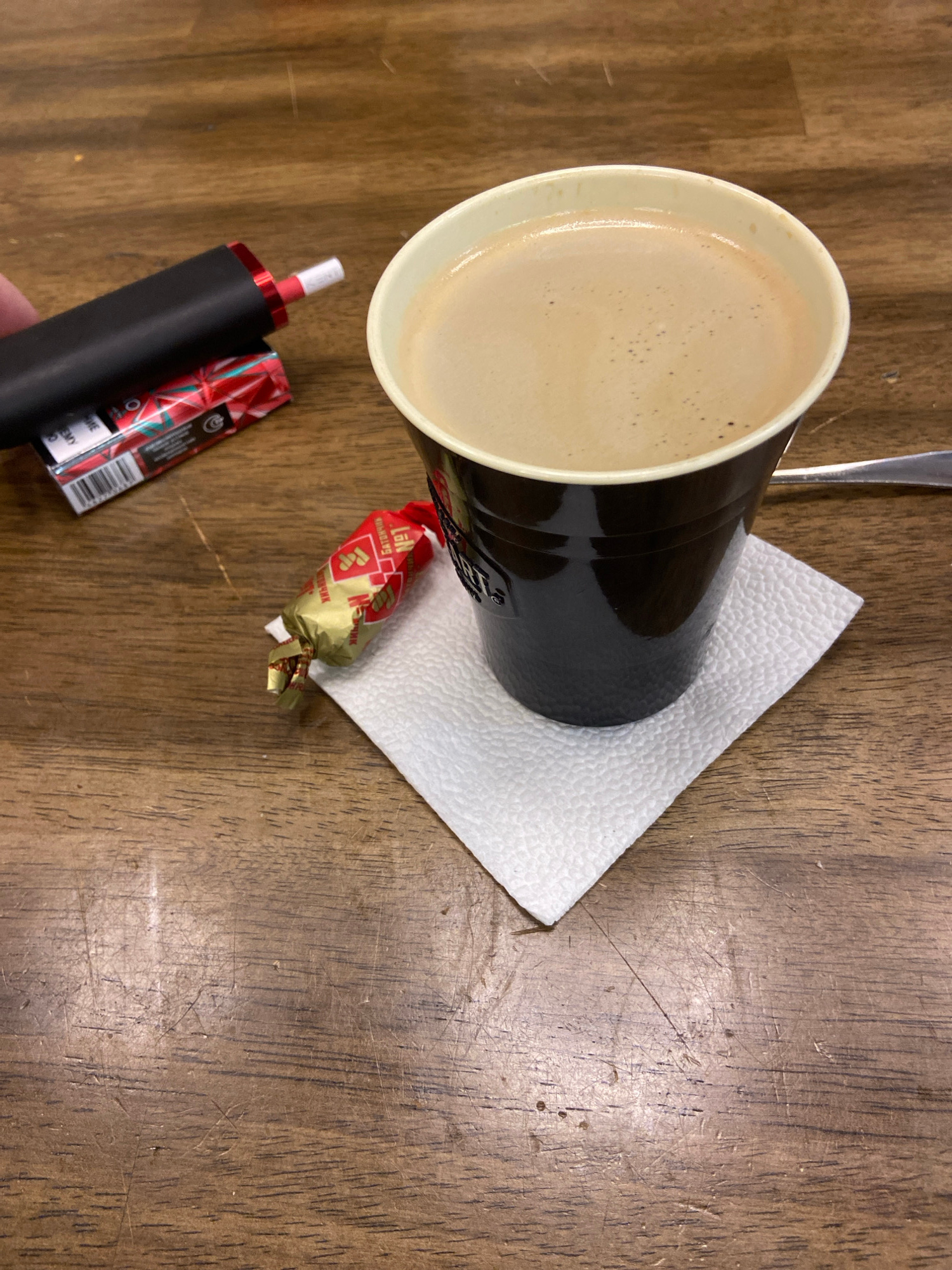 A close-up, eye-level shot shows a coffee cup, a candy, and a small electronic device on a wooden table. The coffee cup is black with a white rim and filled with coffee that has a foamy head. It sits on a white paper napkin. To the left of the cup, a small, individually wrapped candy with red and gold foil is visible. Next to the candy, a black electronic device with red accents is resting on a small package with red and white foil, possibly cigarettes. The wooden table has a visible grain and some scratches. The lighting is neutral, and the overall scene is indoors.