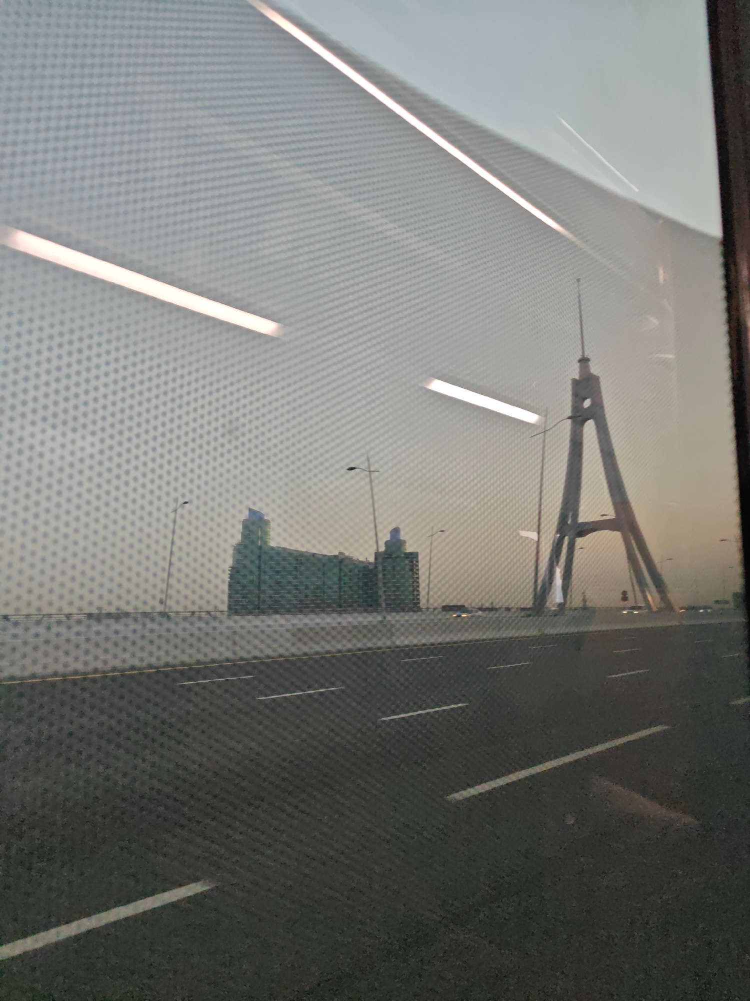 A photograph taken from inside a vehicle, likely on a bridge or elevated highway, shows a view of the Dubai skyline. The foreground is dominated by a road with white lane markings, indicating a highway. In the mid-ground, a large, modern suspension bridge is visible, with its distinctive A-frame towers and cables. The towers are grey and have a geometric, abstract design. Beyond the bridge, a cluster of modern buildings with green facades can be seen, suggesting an urban environment. The sky is hazy and overcast, with the soft light of either dawn or dusk. The image has a slightly blurred quality, possibly due to motion or the texture of the window through which it was taken, which appears to have a fine, dot-like pattern. The overall impression is of a typical, albeit somewhat blurry, view of Dubai's infrastructure and architecture.