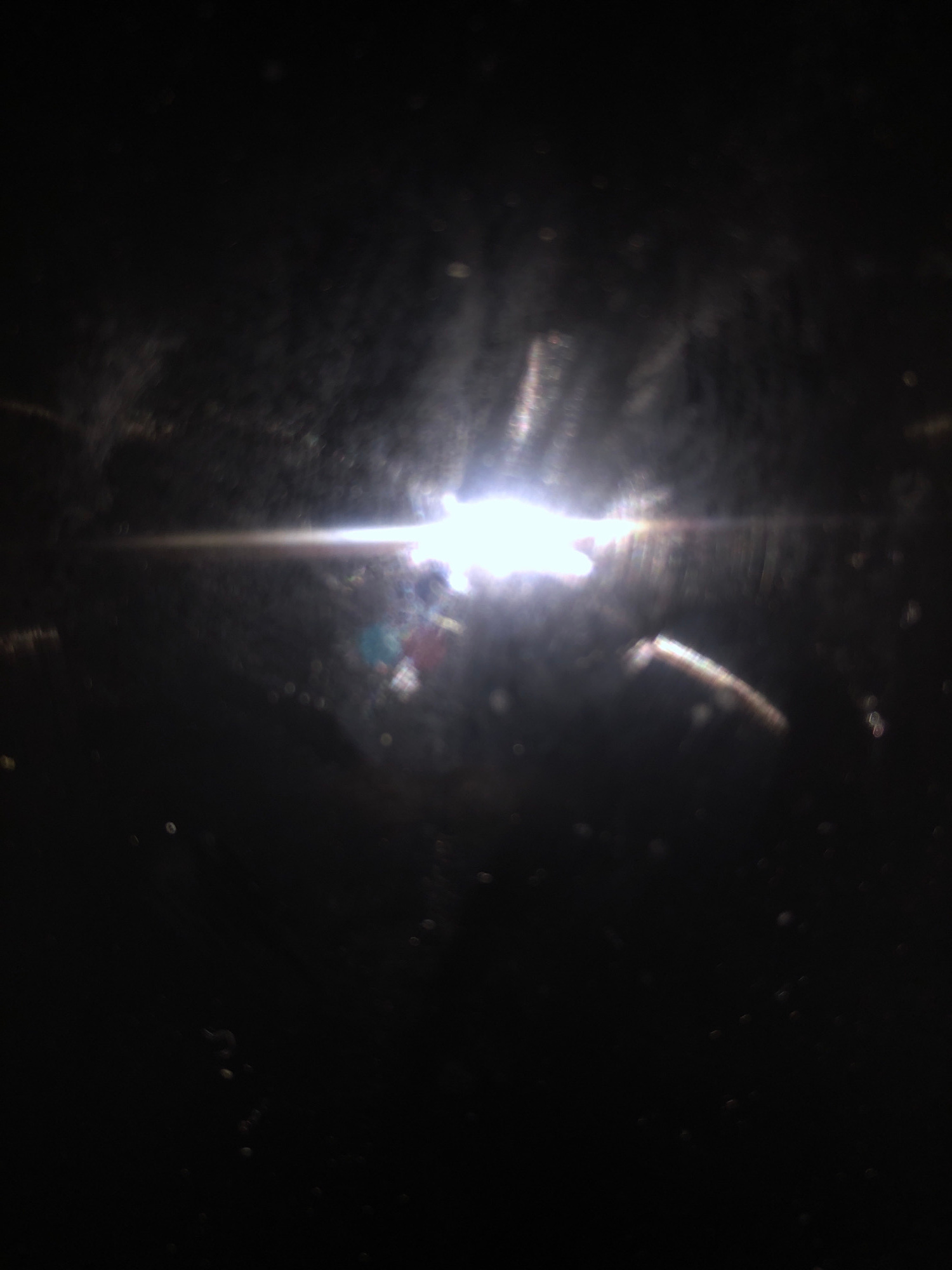 The image shows a bright light source, possibly the sun, shining from the left side of the frame. The light is diffused and creates a lens flare effect, with streaks and a halo of light extending across the image. There are some blurry, out-of-focus shapes and reflections visible in the foreground and background, suggesting the photo was taken through a dirty or textured surface, possibly a window or glass. The overall impression is abstract and dark, with the bright light being the dominant element. There is no discernible location in Jalingo, Nigeria, nor any people or specific objects identifiable.
