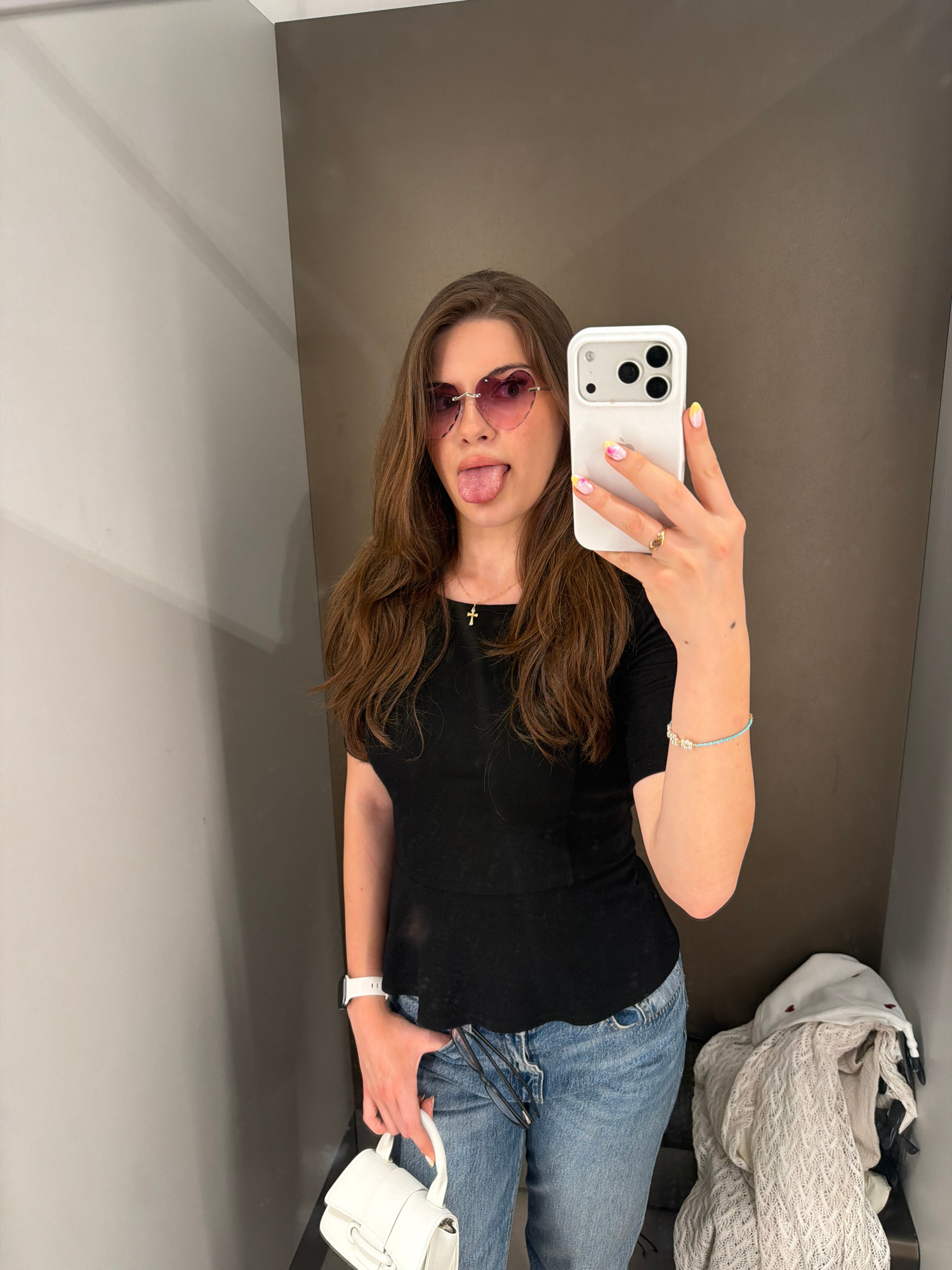 A young woman with long brown hair is taking a mirror selfie in what appears to be a changing room. She is wearing heart-shaped pink sunglasses, a black t-shirt, and light-wash blue jeans. She is sticking her tongue out and holding a white smartphone in her right hand. She has a gold ring on her right ring finger and a beaded bracelet on her right wrist. She is also wearing a white smartwatch on her left wrist and holding a small white purse in her left hand. In the background, there are light gray walls and a darker brown wall. There are also some clothes piled up on the right side of the image, including a white knitted garment. The scene is likely a fitting room within a retail store in the city of Sofia, Bulgaria.