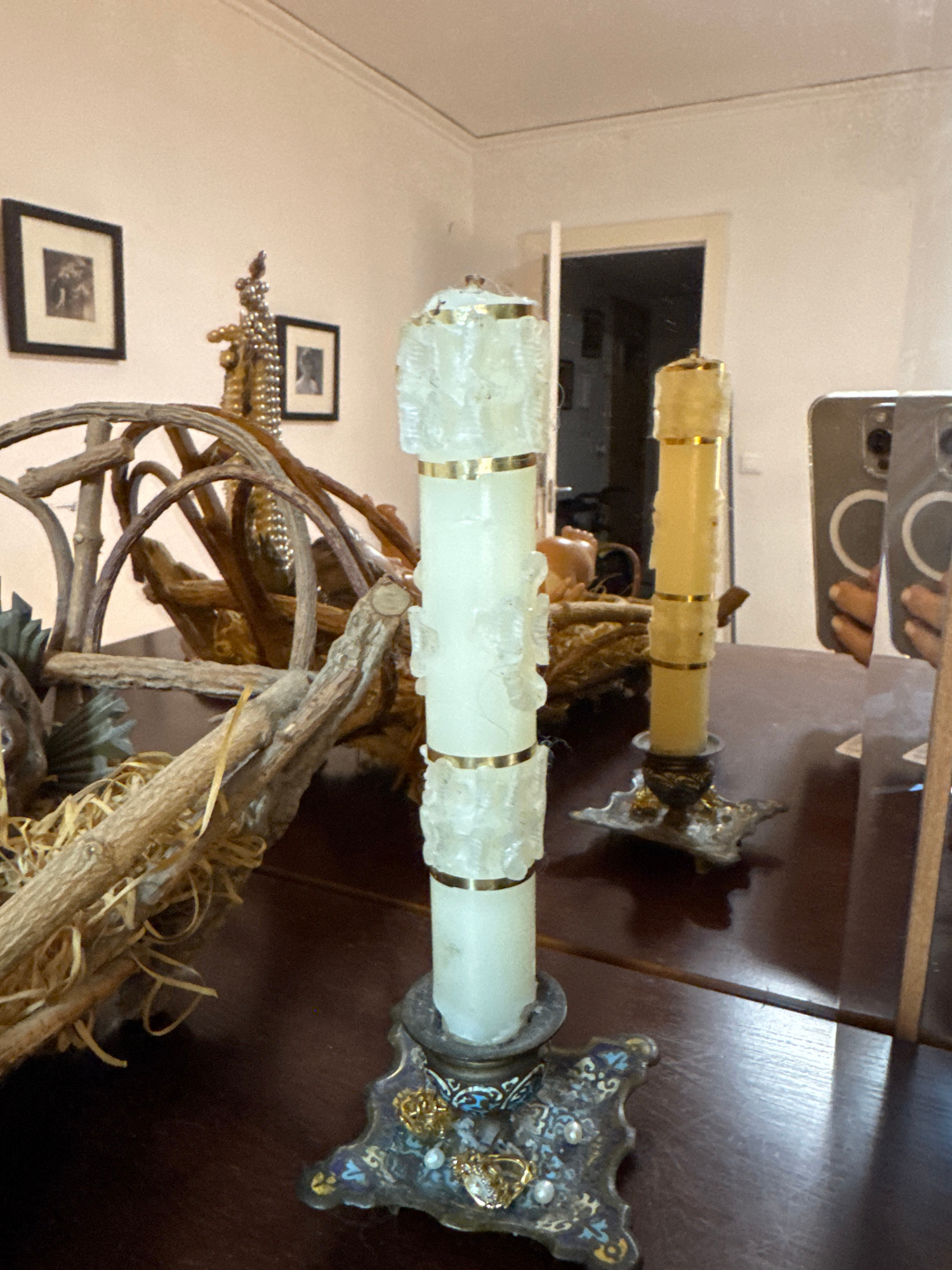 A close-up shot captures an ornate candle holder with a tall, white, ribbed candle featuring gold bands, standing on a dark, reflective surface. To the right, another candle holder with a shorter, yellow candle is visible. The reflective surface, likely a table or dresser, shows a mirrored reflection of the scene, including what appears to be a doorway in the background, suggesting the photograph was taken indoors. Above and behind the candle holders, a decorative wooden structure with branches and straw-like material is present. Two framed pictures hang on a light-colored wall in the upper left corner of the frame. The image is cropped to focus on these elements, implying a domestic setting, potentially in Gandia, Spain, given the context.