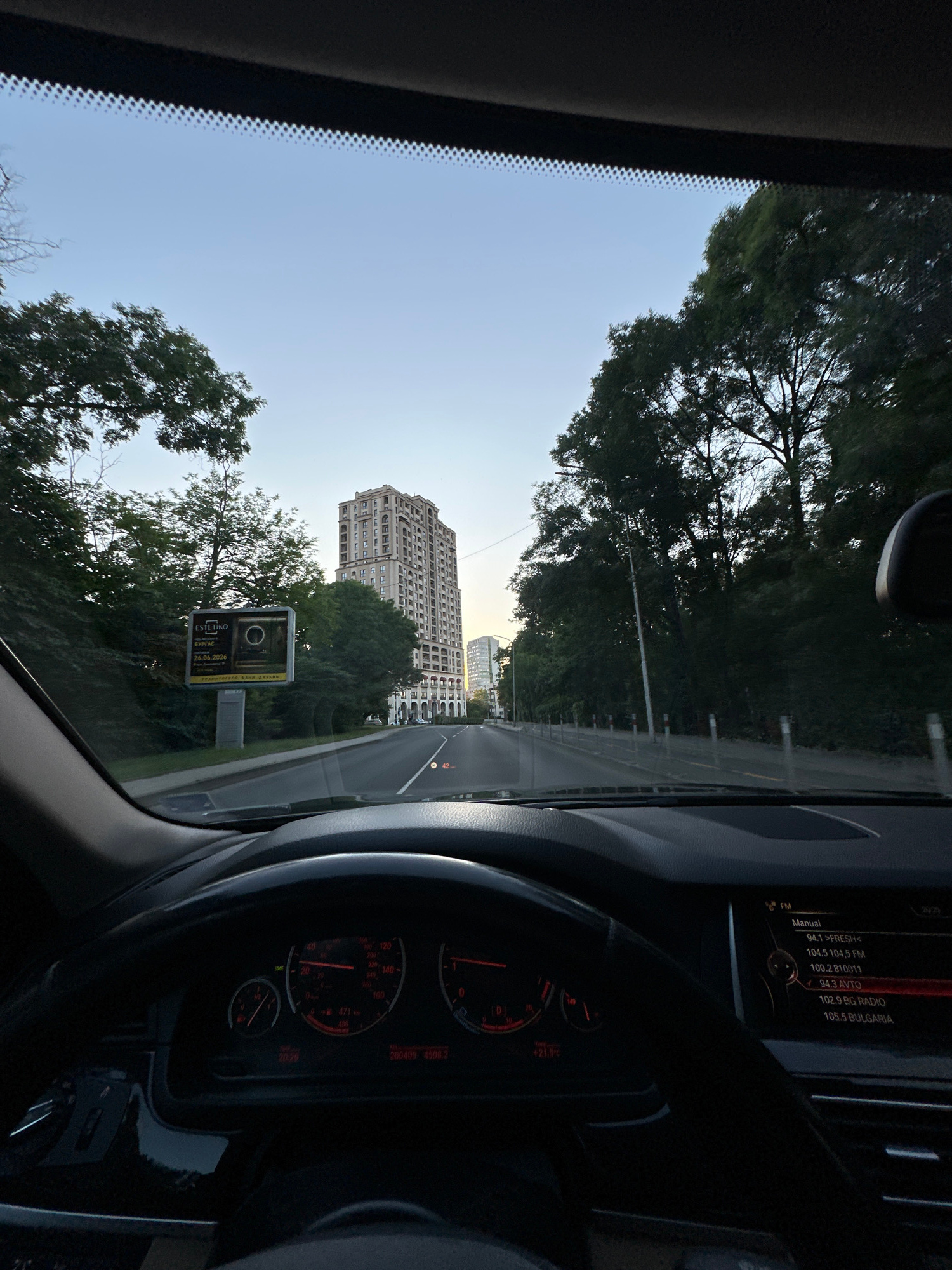 The image is taken from inside a car, looking forward through the windshield. The car is driving on a paved road in a city environment. The road appears to be a multi-lane highway with trees and buildings lining both sides. In the distance, a tall, multi-story building with a beige facade and many windows is visible. To the left of the road, there is a billboard with text and an image. The dashboard of the car is visible in the foreground, including a steering wheel and instrument cluster with red illuminated gauges. The sky above is clear and a light blue, suggesting it is either early morning or late afternoon. The overall scene depicts a drive through an urban area with a mix of nature and architecture.