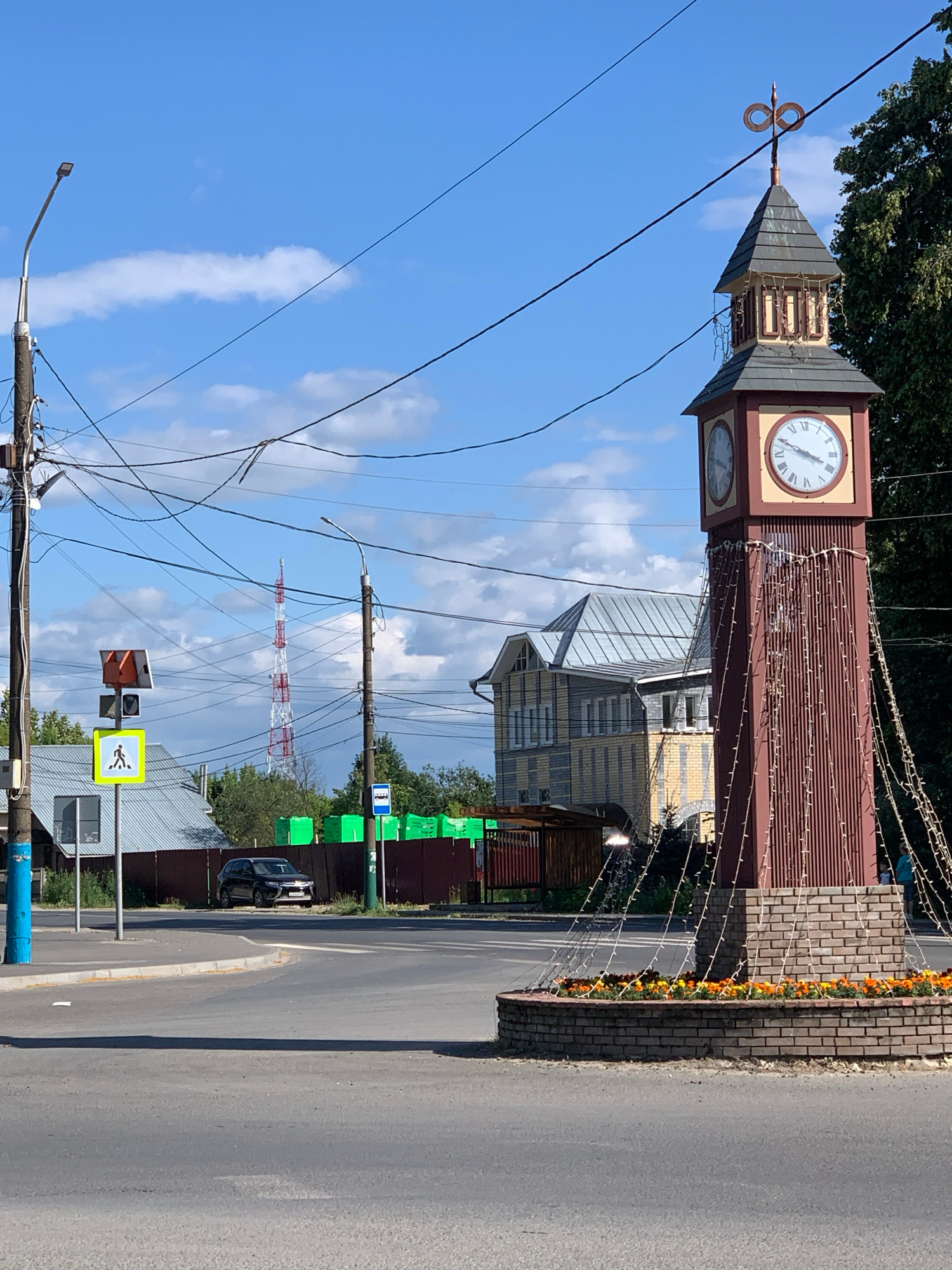 The image depicts an outdoor scene in a town, likely during the daytime under a clear sky with scattered clouds. The primary subject is a tall, reddish-brown clock tower with a peaked roof, adorned with decorative lights that appear to be turned on. The clock face is visible, showing Roman numerals and hands indicating a time. Next to the clock tower, a brick wall with a row of orange flowers runs along a paved road.
In the background, several buildings with distinctive architectural styles, including a yellow and grey building, are visible. A tall, red and white transmission tower stands in the distance. The scene is a mix of urban infrastructure and natural elements, with trees visible behind the buildings.
Several traffic signs and utility poles with numerous wires are also present, suggesting a busy street or intersection. One sign is a pedestrian crossing signal, another is a bus stop sign, and there are green rectangular signs on poles. A dark-colored car is parked on the side of the road. The overall impression is a functional, perhaps slightly aged, town setting on a bright day. There are no people or animals clearly visible in the image.
The location is identified as Pavlovo, Russia, which provides a geographical context for the scene.