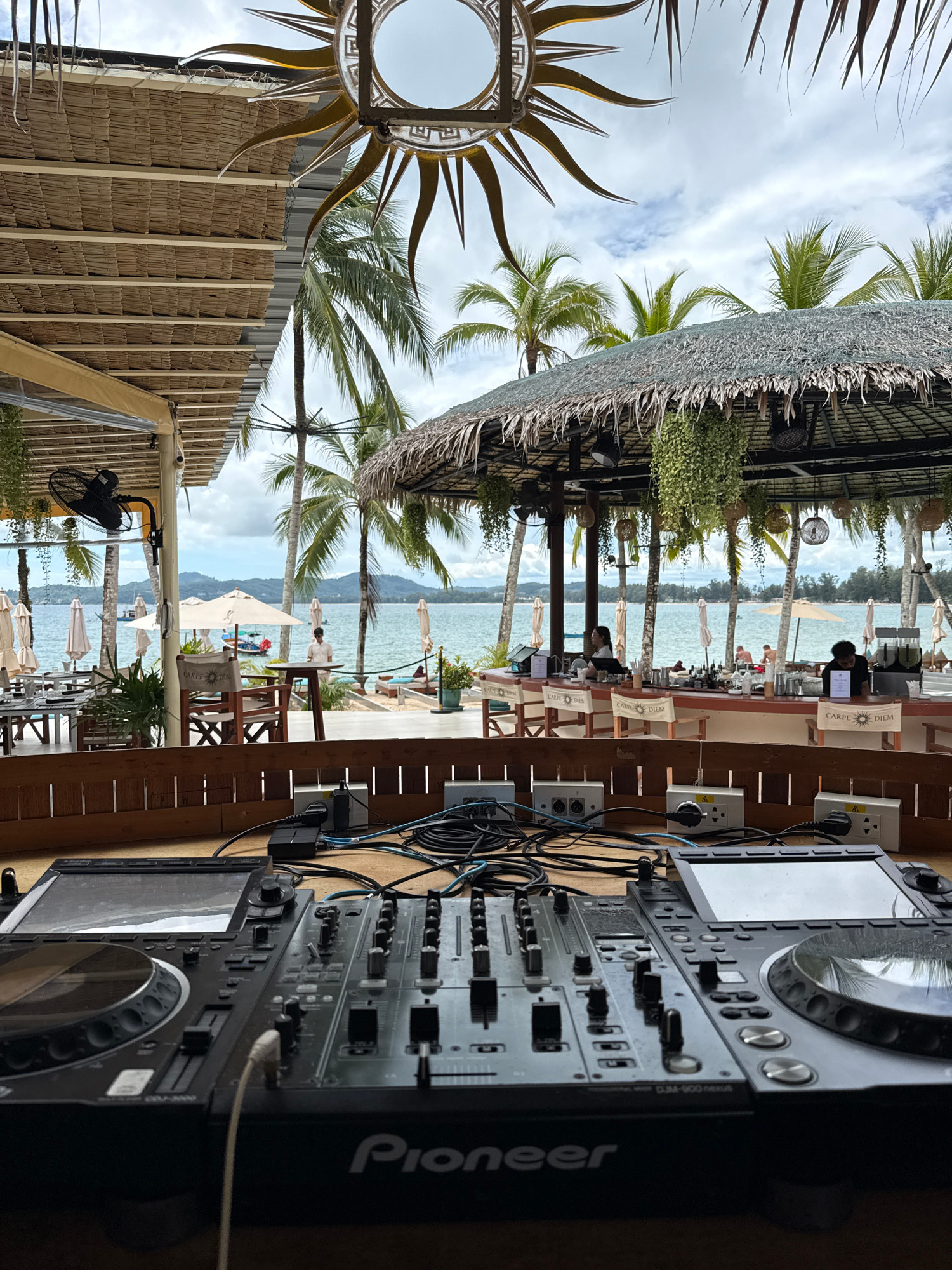 The image captures a view from behind a DJ setup, looking out onto a beachside bar and restaurant. In the foreground, a Pioneer DJ controller and mixer are visible, with various cables and plugs connected to power outlets. Beyond the DJ equipment, a thatched-roof bar area is set up with seating and tables. Several people are present at the bar, and some are seated at tables. The background shows a calm ocean with a few boats, and a treeline with palm trees. Beach umbrellas and lounge chairs are also scattered along the shoreline. The sky is overcast with some visible clouds. The overall setting appears to be a tropical beach resort.