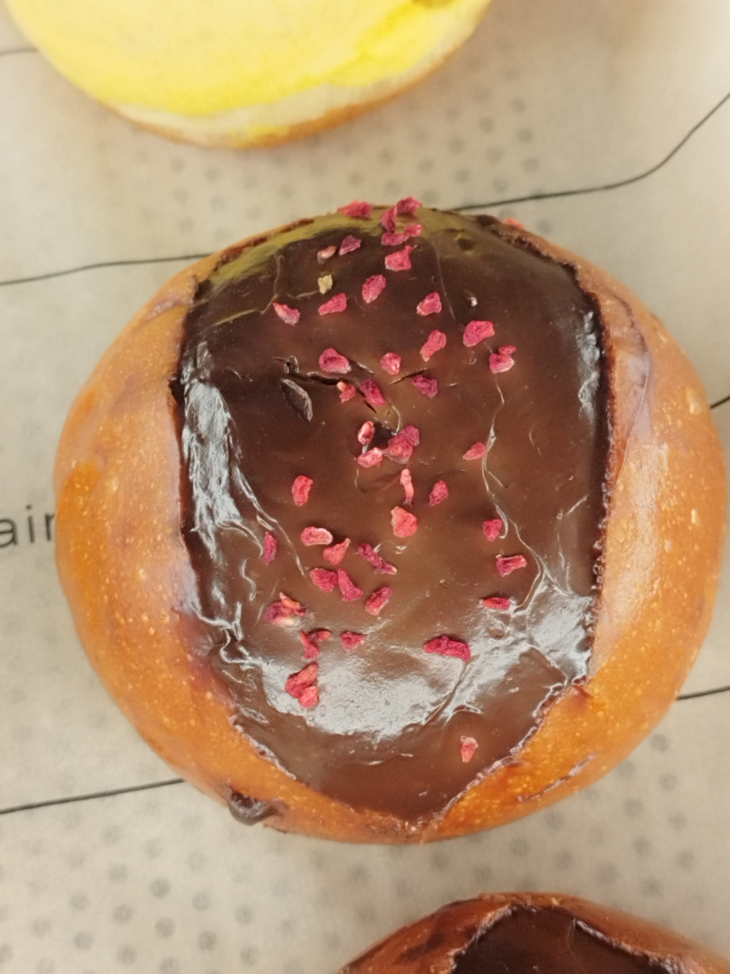 A close-up, high-angle shot captures a chocolate-glazed pastry adorned with small, dried red pieces, likely fruit. The pastry has a golden-brown, slightly irregular crust and sits on a light-colored surface with a dotted pattern. To the upper left, a portion of another pastry with a yellow glaze is visible. The background includes faint text, potentially indicating a location or brand. The overall setting appears to be indoors, possibly a bakery or food display, without any discernible connection to Saint Petersburg, Russia.
