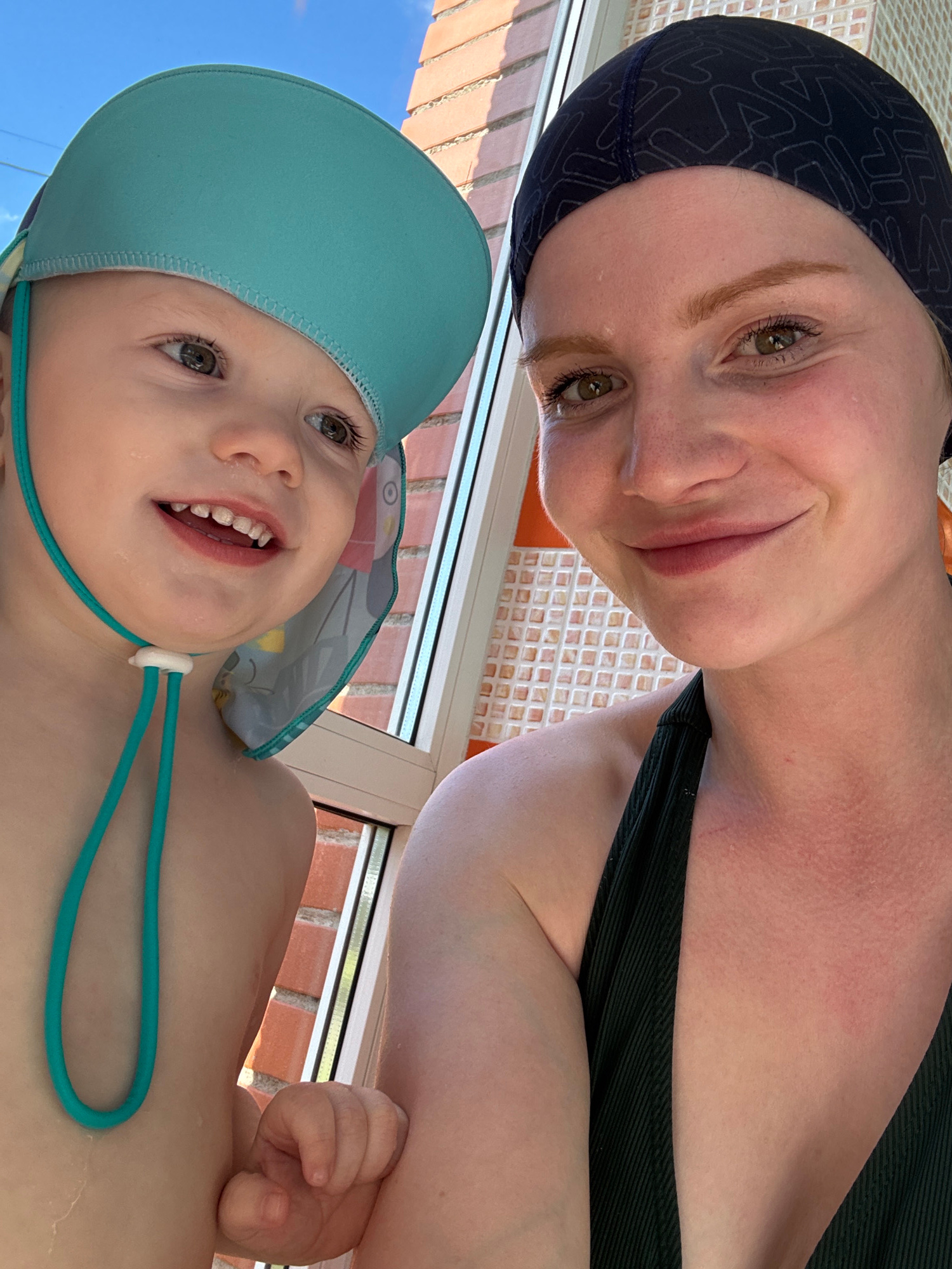 This is a close-up, vertically oriented selfie taken outdoors. The image features two subjects: a woman and a baby. The woman is in the upper half of the frame, wearing a dark swim cap with a repeating geometric pattern. She has light skin and her eyes are green. She is smiling and looking directly at the camera. Her neck and shoulders are visible, and she is wearing a dark, ribbed garment, likely a swimsuit.
Below the woman, the baby occupies the lower half of the frame. The baby is wearing a light blue swim hat with a wide brim and a drawstring. The baby has large, expressive green eyes and is smiling widely, showing teeth. Small water droplets are visible on the baby's face and chin.
The background suggests an aquatic setting. Behind the subjects, a large window or glass partition reveals a brick wall and the blue sky, indicating it might be daytime and possibly sunny. The presence of swim caps and the aquatic theme suggest the activity taking place is swimming or a related water activity, likely at an indoor pool with an outdoor view or an outdoor pool. The overall mood is joyful and happy, conveyed by the smiles of both individuals. No visible text is present in the image. The location context is Saint Petersburg, Russia.