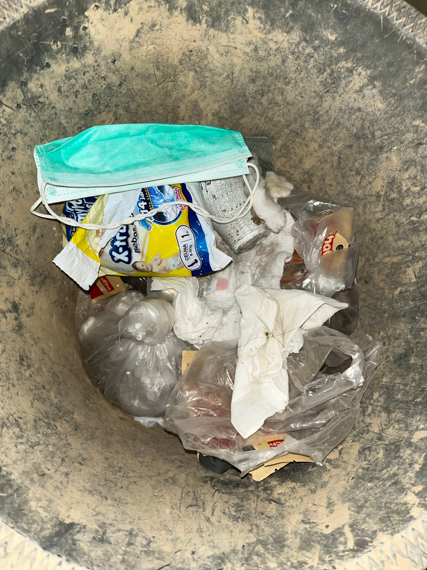 A close-up, top-down view inside a weathered, dark gray trash bin filled with various discarded items. A light blue surgical mask rests on top of a yellow and white plastic package, which appears to be baby diapers, featuring an image of a baby. To the right of the diaper packaging, a crumpled silver can and several white paper tissues are visible. The bin also contains other plastic-wrapped items, some appearing to be food packaging with red and yellow labels, and transparent plastic bags holding more unidentifiable contents. The interior of the bin is coated with a layer of dust and dirt, suggesting it is located outdoors or in a dusty environment. The exterior of the bin shows signs of wear and tear. The scene is set in the city of Kota Kuala Simpang, Indonesia.