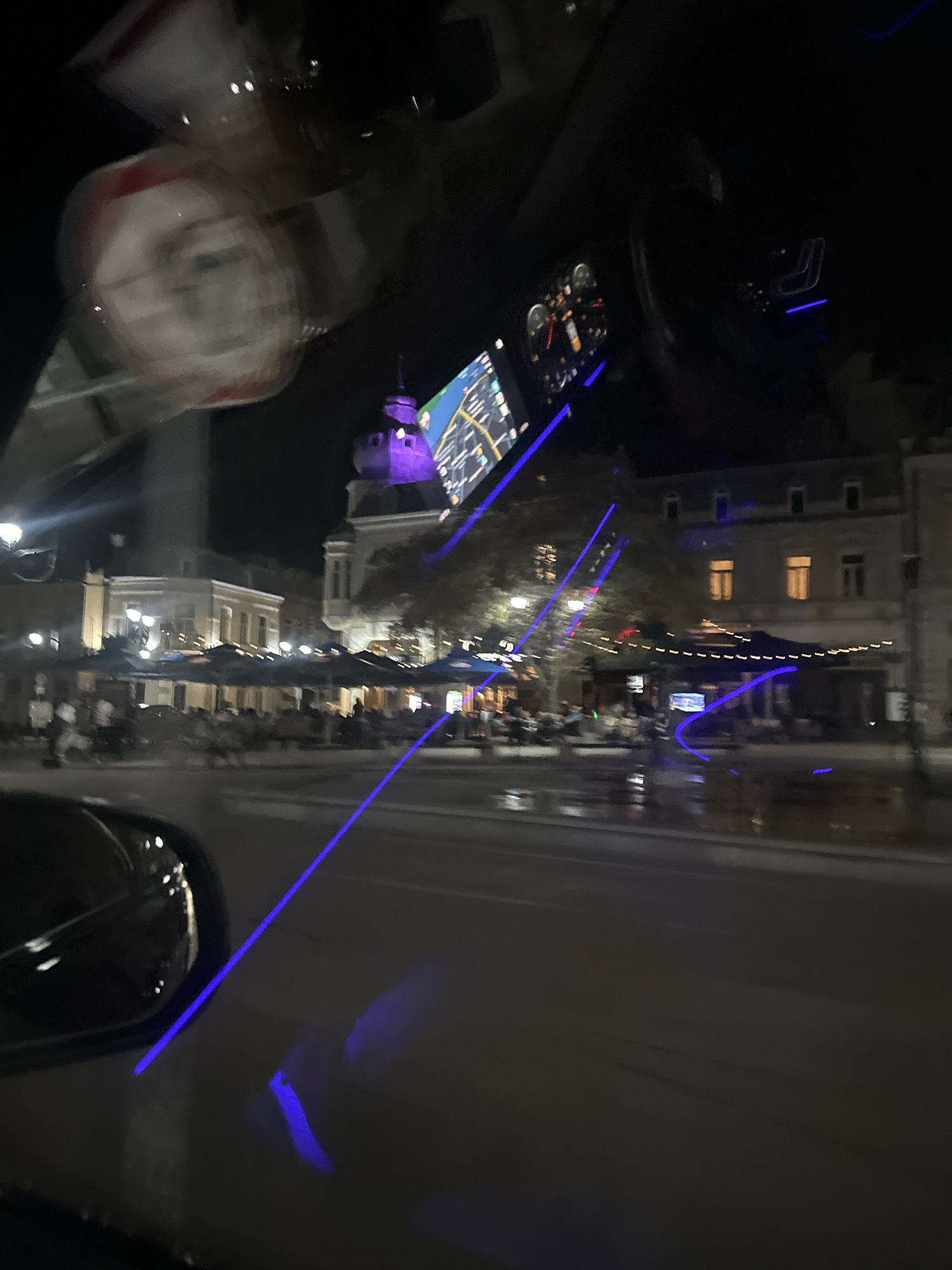 The image captures a night scene, likely from inside a vehicle. The perspective is skewed, suggesting the photo was taken while moving. In the foreground, a blurry object, possibly a drink container, is visible on the left. In the center, a car's dashboard or console is illuminated, displaying a GPS navigation screen with a map. The background reveals a street scene at night, with buildings lining the road. Streetlights and lights from within the buildings create a warm glow. Blue light strips are prominently visible, extending across the scene, likely part of the vehicle's interior lighting or decorative elements. There are reflections on the surfaces, indicating a glossy finish. The overall impression is one of travel at night through an urban environment. The presence of buildings with illuminated windows suggests a populated area. The blurred nature of the image and the blue light accents create a dynamic and somewhat futuristic feel. Without further context, it's difficult to determine the exact activity or event, but it strongly suggests a journey. The darkness and the artificial lighting indicate it is nighttime.