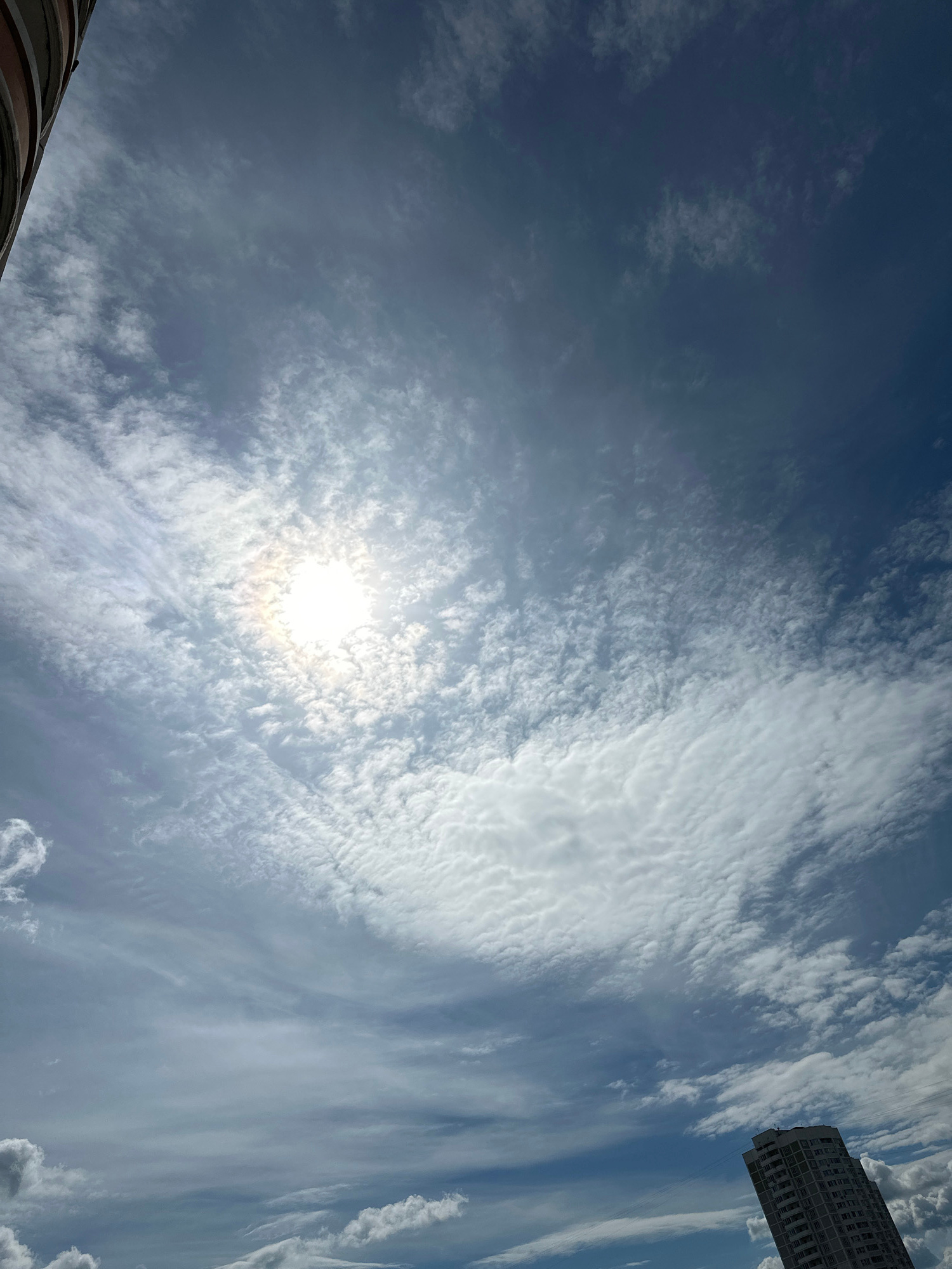 A low-angle view captures a bright, partially obscured sun illuminating a sky filled with scattered white and gray clouds. The sun's rays create a halo effect through the thin cloud cover. In the upper left corner, a curved, textured architectural element, possibly part of a building, is visible. In the lower right quadrant, the top section of a tall, modern building with many windows is depicted. The overall scene is an urban sky with elements of architecture and weather. The location is Podolsk, Russia.