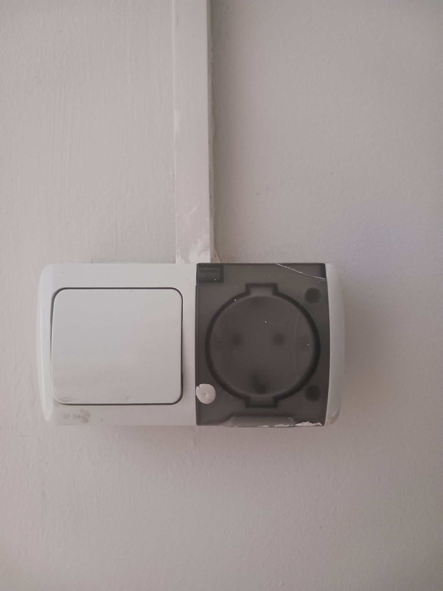 In an indoor environment, against a plain white wall with a thin vertical white conduit, a white electrical outlet with a grey transparent protective cover is mounted. To the left of the outlet is a white light switch. The outlet cover appears to be slightly scratched. Below the light switch, faint lettering that resembles "IP 54" is visible. The scene is brightly lit, and there are no people present.