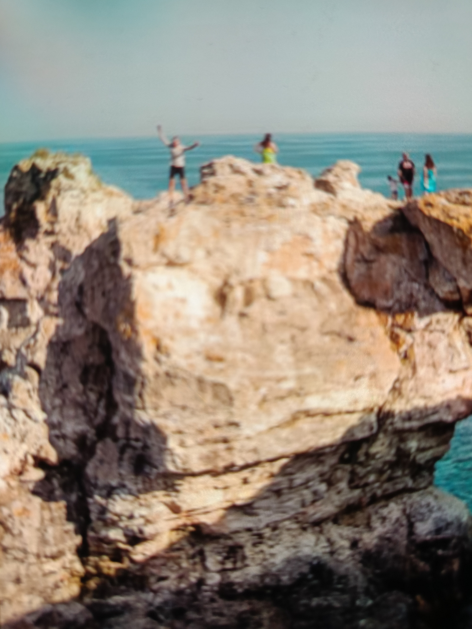 The image shows a group of people standing on a large rock formation overlooking the sea. The rock is sandy and appears to be part of a cliff face. The sea is blue and calm, and the sky is clear and bright. There are several individuals visible on the rock, appearing to be enjoying the view and the outdoors. One person is standing with their arms raised in the air, appearing celebratory or excited. Others are standing in groups or individually. The location is identified as Varna, Bulgaria. The overall scene depicts a recreational activity in a coastal environment.
