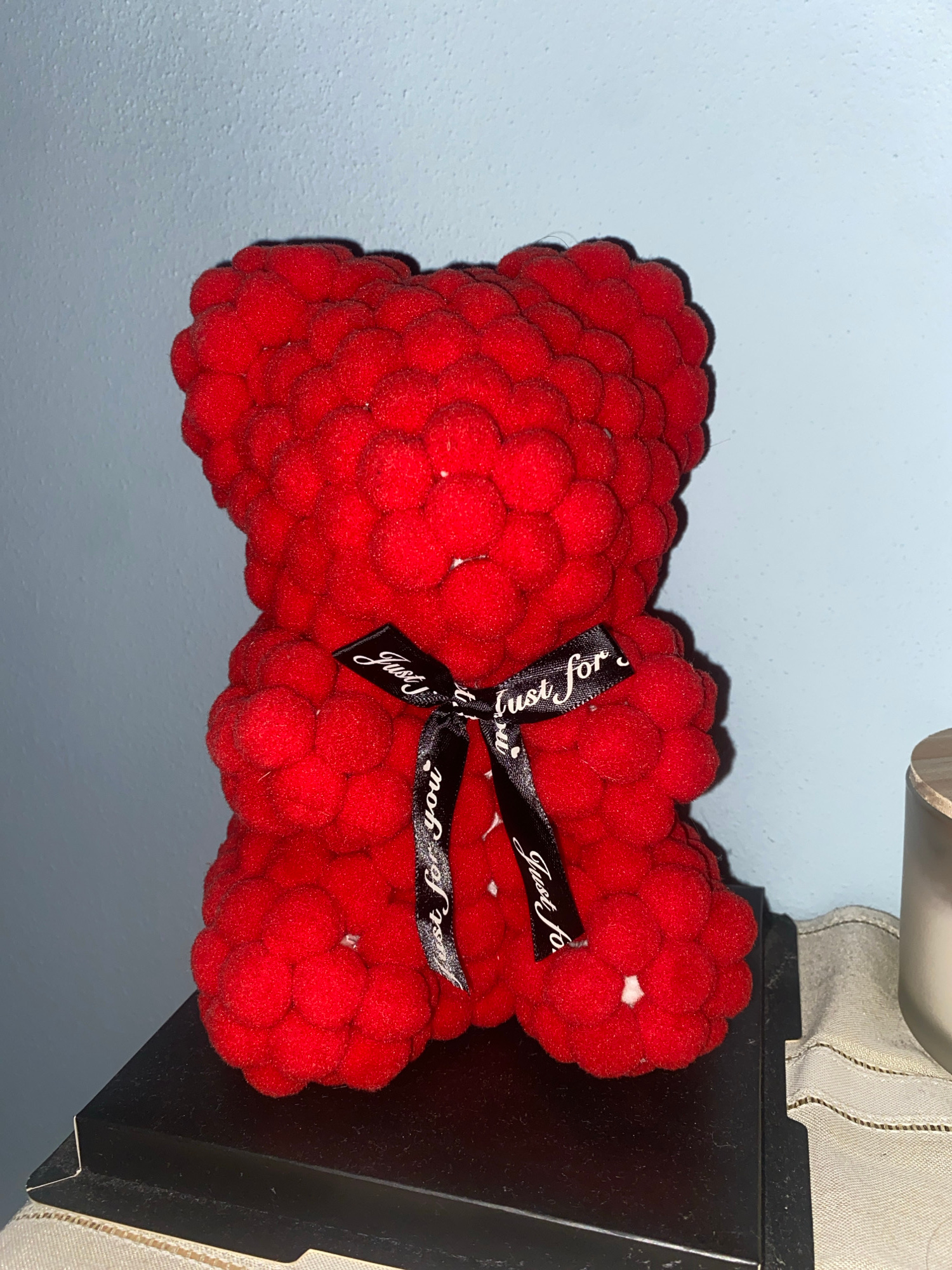The image shows a red rose bear ornament with a black ribbon that says "Just for you" in white lettering. The bear is sitting on a black surface, which appears to be a box or stand. Behind the bear is a light blue wall. To the right of the bear, part of a candle is visible. The scene is indoors and well-lit, with the light source appearing to be from the front and slightly to the left. The image is a close-up shot, focusing on the bear ornament. There is no indication of the city of Brescia, Italy, or any other specific location.