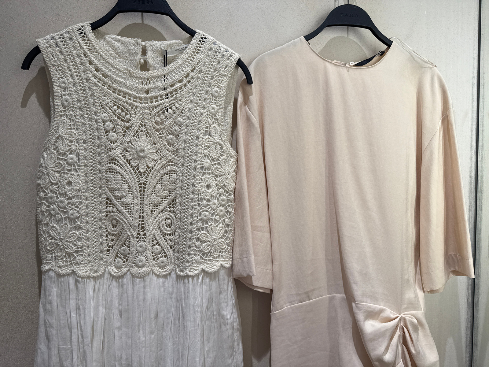 This is a medium shot of two dresses hanging on a wall, likely in a retail setting. The dress on the left is white and features intricate crocheted detailing on the bodice, with a delicate floral pattern. The lower half of this dress is made of a pleated fabric, creating a flowy silhouette. The dress on the right is a pale peach or light beige color and appears to be made of a smooth, satiny material. It has a relaxed fit with wide sleeves and a draped detail at the hem, tied into a knot. Both dresses are on black hangers. The background is a plain, light-colored wall, suggesting an indoor environment. The lighting is even, indicating it is likely daytime or under artificial store lighting. The brands "ZARA" are visible on the hangers of both dresses. The scene is static, with no people or activity other than the display of the garments.