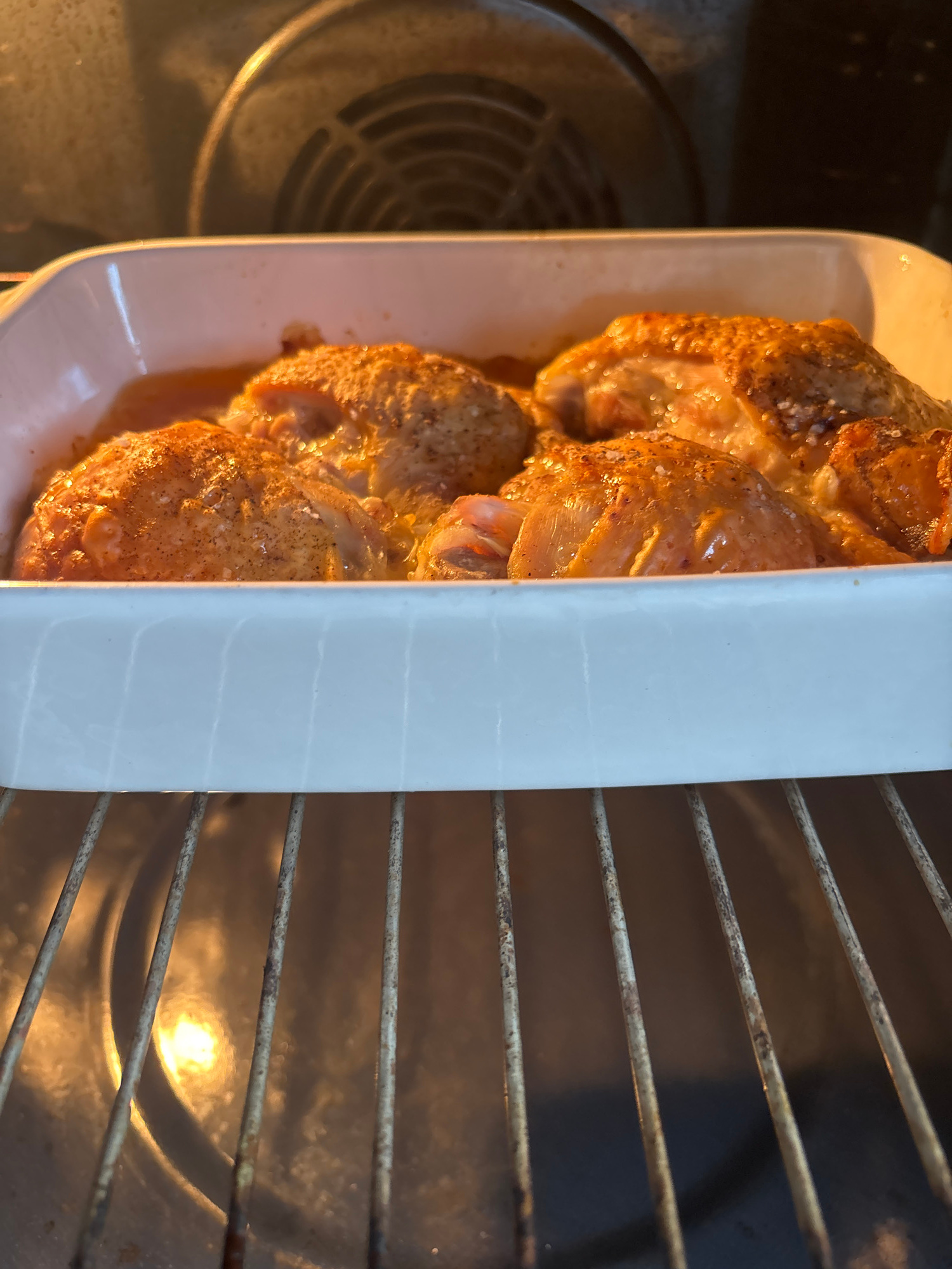 A close-up, overhead shot shows chicken thighs baking in a white rectangular baking dish, placed inside an oven. The chicken pieces are browned and seasoned with visible salt crystals and spices. The baking dish is positioned on a wire rack within the oven, with other oven racks visible in the foreground and background, suggesting the dish is on a middle or lower rack. The oven interior is lit, with a circular heating element or fan visible on the left side of the image. The overall scene is one of home cooking. There is no visible text in the image. The context provided indicates the location is Bila Tserkva, Ukraine, but this is not discernible from the image itself.