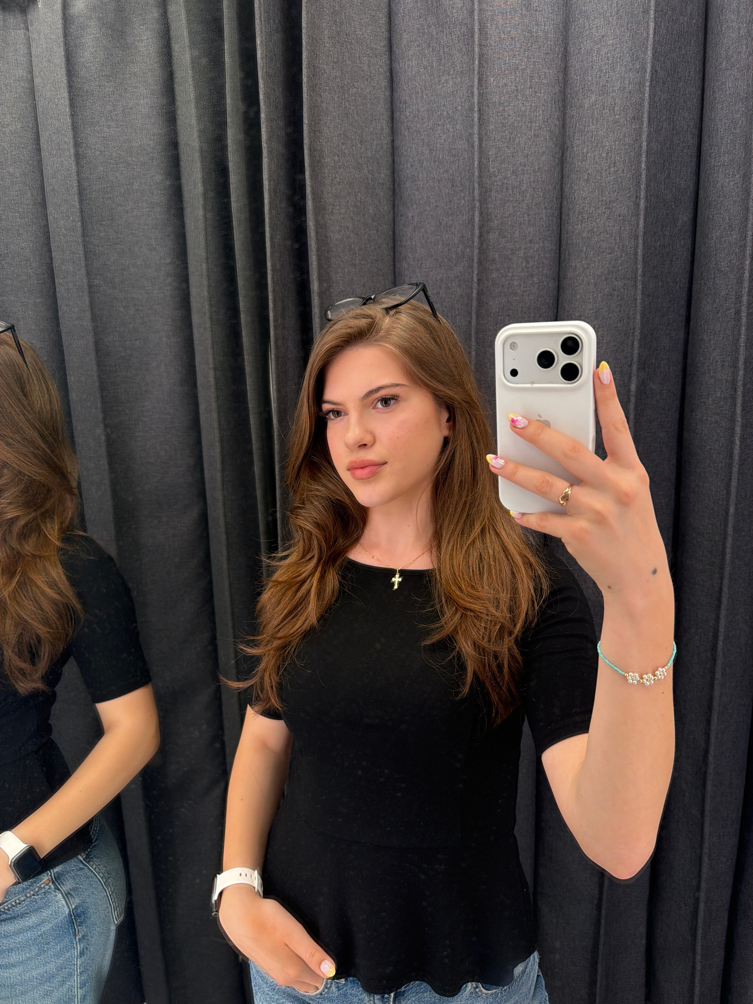 In a room with dark gray pleated curtains, a young woman with long, wavy brown hair takes a selfie in a mirror. She is wearing a black short-sleeved shirt, blue jeans, a white smartwatch on her left wrist, and a thin gold necklace with a cross pendant. Her right hand, holding a white iPhone, is adorned with a gold ring and a turquoise beaded bracelet with silver flower charms. Her fingernails are painted with a colorful design. A reflection of the woman is visible in the mirror to her left, also wearing a black shirt and blue jeans, with a white smartwatch on her left wrist. The scene appears to be in a changing room or fitting room, possibly in a retail establishment in Sofia, Bulgaria.
