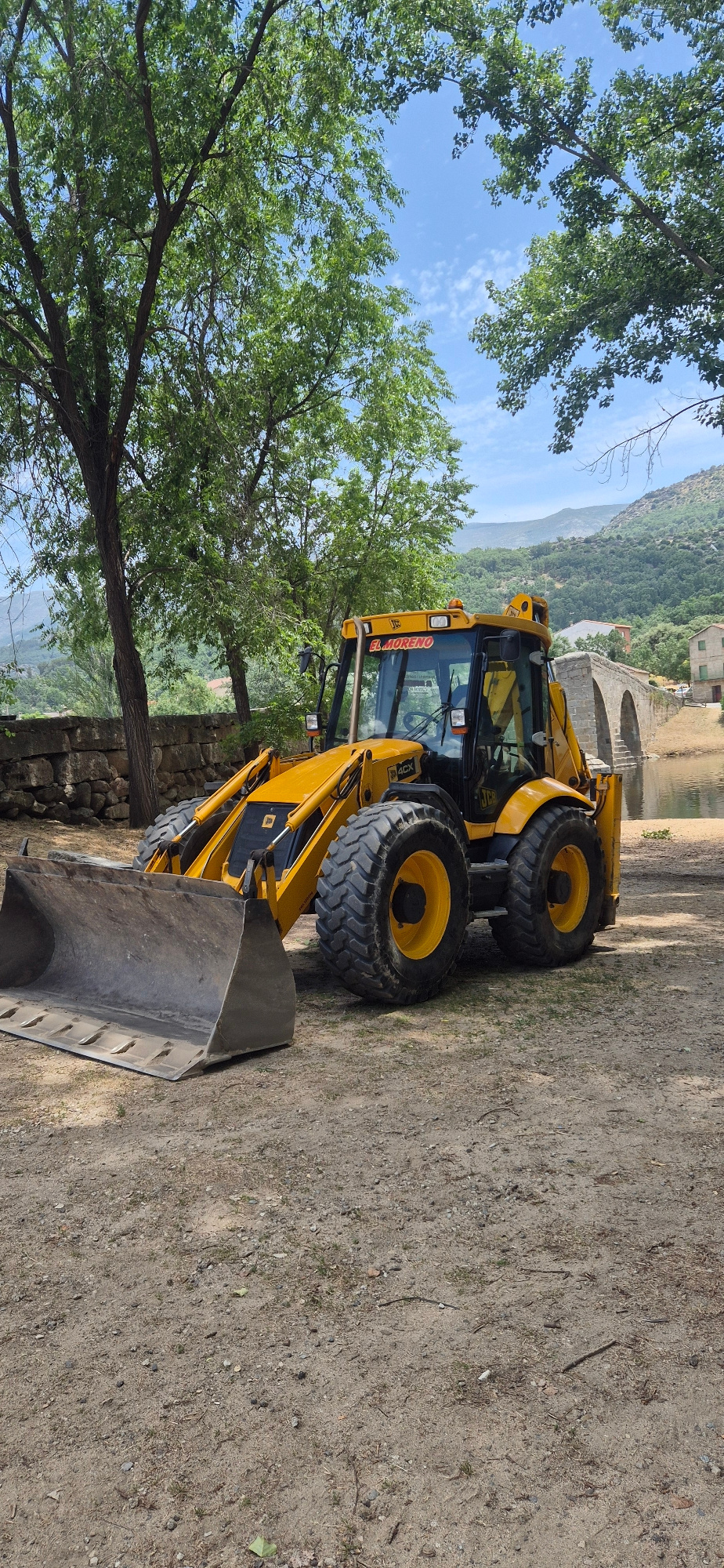 A bright yellow JCB backhoe loader is parked on a dirt path. The machine has a large bucket at the front and large tires. The cab of the backhoe has "EL MORENO" written on it in red letters. In the background, there is a stone bridge with multiple arches spanning a body of water. Trees with green leaves are on either side of the scene, and hills covered in trees can be seen in the distance under a blue sky with scattered clouds. The ground is a mix of dirt, small stones, and some sparse green vegetation.