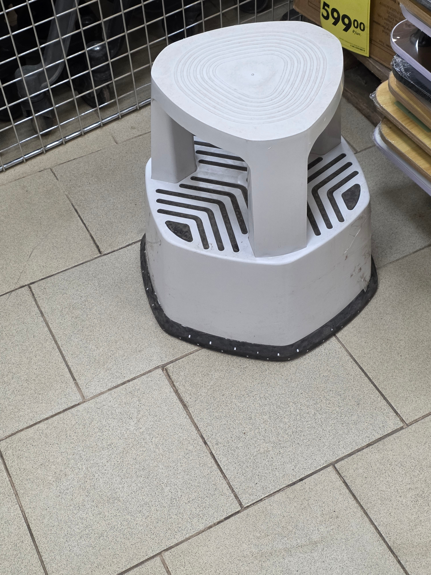 A white, two-tiered plastic step stool with a textured top surface is positioned on a light beige tiled floor. The stool has a wider base with slots on its upper surface and a narrower top. A black rubber trim runs around the base of the stool. To the left of the stool, a metal wire cage or shelving unit is visible. To the right, a yellow price tag with black lettering reading "599.00" and a stack of items, possibly skateboards or longboards, are partially visible. The scene appears to be in an indoor retail environment.
