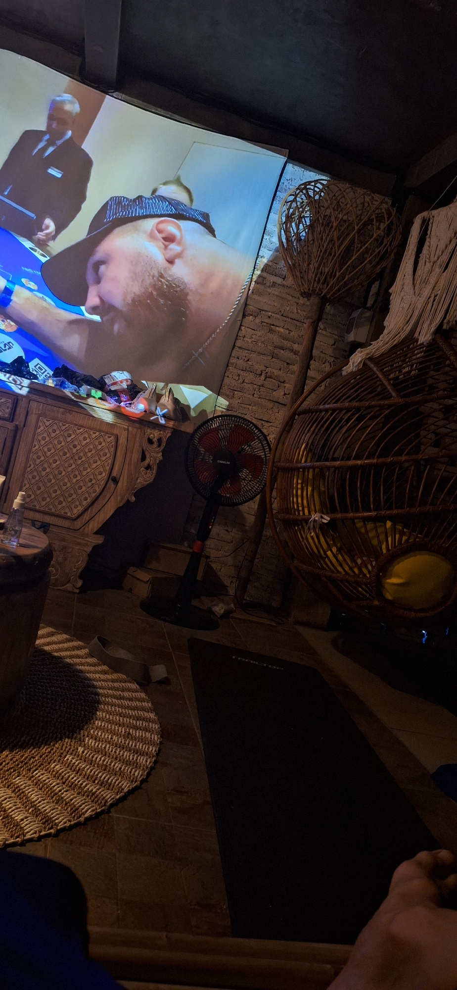 An interior scene shows a large projection screen displaying a man wearing a hat and a partially visible man in a suit. In the foreground, a wooden cabinet with ornate carvings holds various items, including a bottle and a fan. To the right, a woven hanging chair is visible, and a brick wall forms the backdrop. A circular woven mat is on the floor, along with a black mat. The scene appears to be in a dimly lit room with a relaxed, possibly entertainment-focused atmosphere. This image was captured in Kuta Selatan, Indonesia.