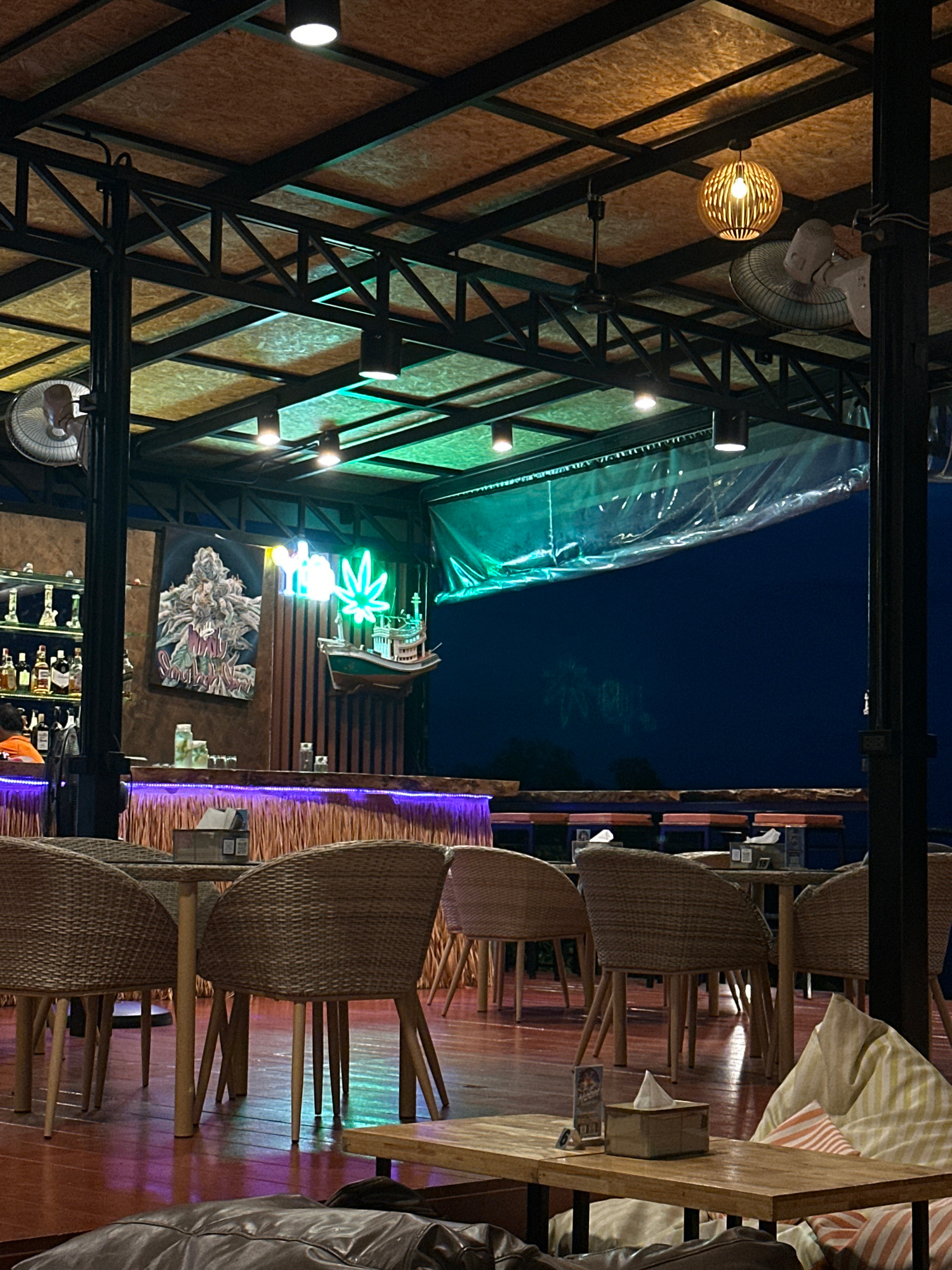 The image captures a nighttime scene inside a dimly lit establishment, possibly a bar or restaurant, with a rustic and tropical ambiance. The camera is positioned at an angle, looking upwards and sideways, giving a unique perspective of the interior.
In the foreground, several woven chairs surround wooden tables. The chairs are a prominent feature, their intricate weaving suggesting a handcrafted or natural material. Some of these chairs are empty, while others appear occupied, though no specific individuals are clearly identifiable. Cushions with striped patterns are visible on some of the seats, adding a touch of color and comfort.
The setting is characterized by exposed structural beams and wooden paneling on the walls, creating an open, airy feel, even with the ceiling overhead. Lighting plays a significant role in the atmosphere, with warm, yellow light emanating from a decorative woven pendant lamp, and spotlights casting bright beams on the wooden walls. In the background, a neon sign shaped like a marijuana leaf glows brightly in green, accompanied by other illuminated elements. Below the neon sign, a framed artwork depicts what appears to be a cannabis bud. Further back, shelves lined with bottles suggest a bar area.
The time of day is clearly nighttime, indicated by the dark sky visible through an opening in the structure, and the artificial lighting within the establishment. The overall mood is relaxed and inviting, with the warm lighting and natural materials contributing to a cozy atmosphere. The presence of the cannabis leaf imagery suggests a potential theme or specific type of establishment.
Visible text includes the words "BIG BUD" written on the artwork below the neon sign. The location context provided is Baan Bang Rak, Thailand, which aligns with the tropical aesthetic hinted at by the materials and design elements.