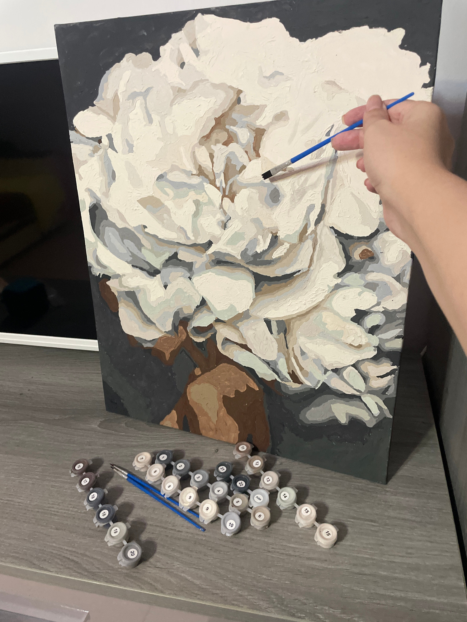 The image depicts a paint-by-numbers project in progress, featuring a large white flower with muted tones of cream, grey, and brown against a dark background. A hand holding a blue-handled paintbrush is actively applying paint to the canvas, specifically to the flower's petals. In the foreground, arranged on a wooden surface, are numerous small containers of numbered paint pots, also in shades of cream, grey, and brown, alongside two blue-handled paintbrushes. The setting appears to be indoors, on a grey wooden desk or shelf, with a portion of a television visible in the background. There is no information in the image to suggest a location in Sochi, Russia.