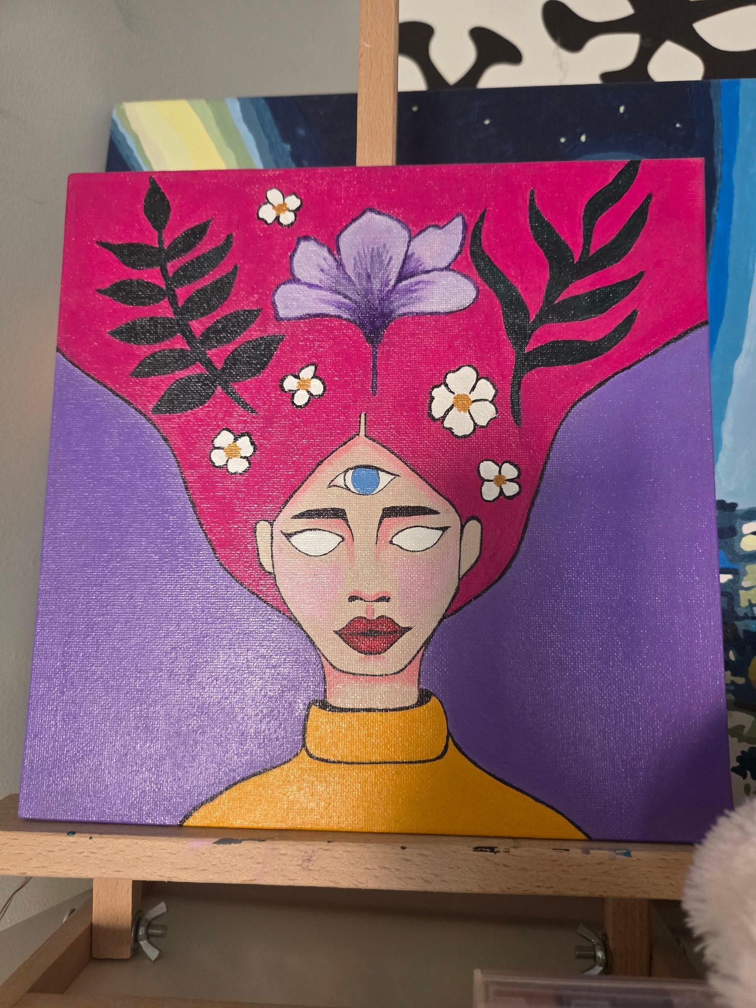 The image is a medium shot of an acrylic painting of a woman with stylized features. The painting is displayed on a wooden easel. The woman has pink hair that flows into a purple background. She has a third eye on her forehead, with blue iris. White flowers and black leaf-like shapes are embedded in her pink hair. She wears a yellow turtleneck. The painting's style is flat and graphic, with bold outlines. In the background, other paintings are partially visible, suggesting an art studio or exhibition setting. One painting to the left has abstract bands of color, and another to the right depicts a cityscape at night. The location of Brzeg, Poland, is not discernible from the image.