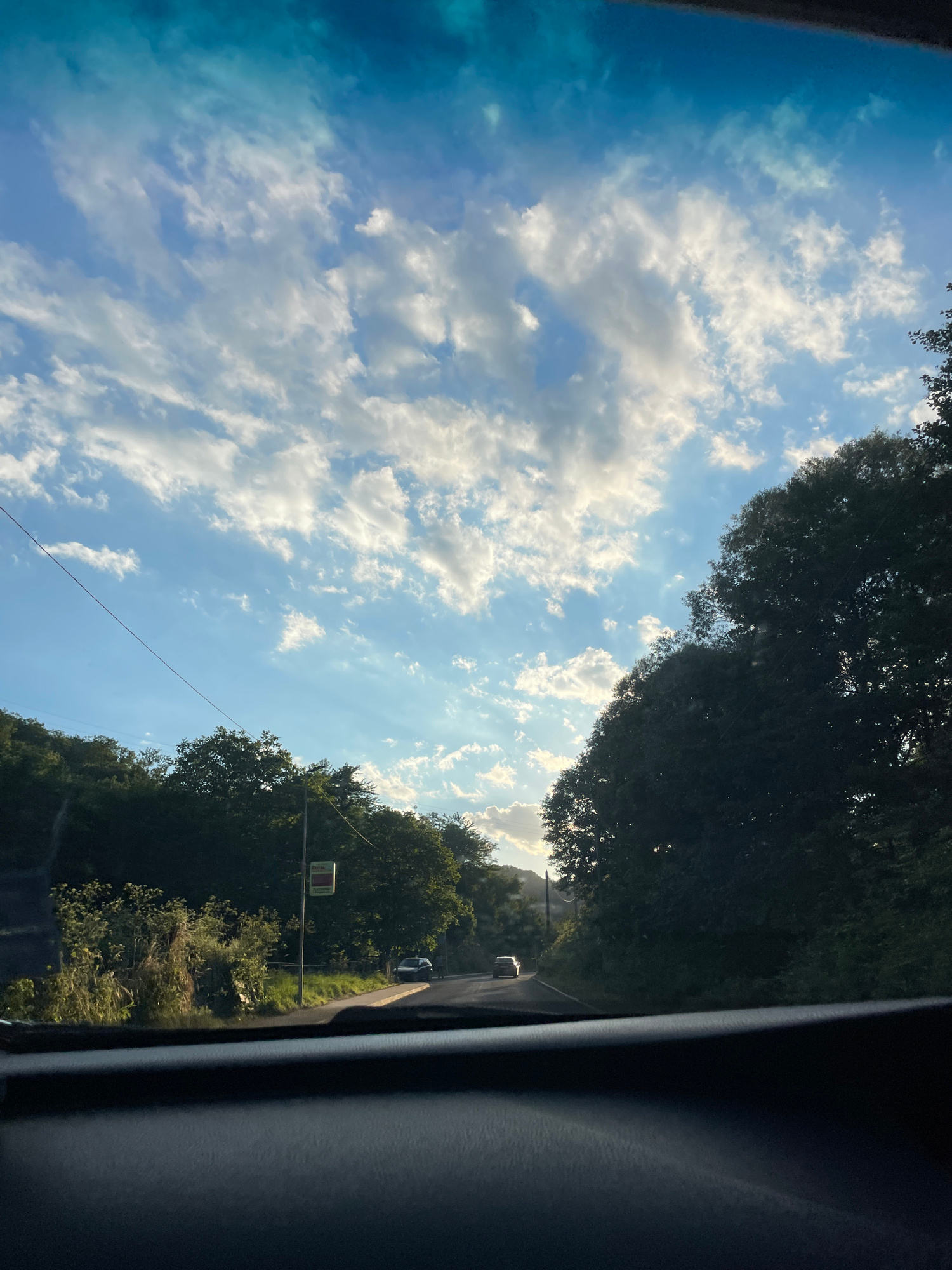 The image shows a view from inside a car looking out through the windshield at a road winding through a lush, green landscape. The sky is a vibrant blue, dotted with fluffy white clouds that are illuminated by the setting sun, casting rays of light through the atmosphere. Tall trees line both sides of the road, creating a natural canopy. A few cars are visible on the road ahead, suggesting this is a public route. The dashboard of the car is in the foreground, obscuring the bottom portion of the view. The overall impression is one of a peaceful drive through a rural area. The location is identified as being in the city of Zlatograd, Bulgaria.