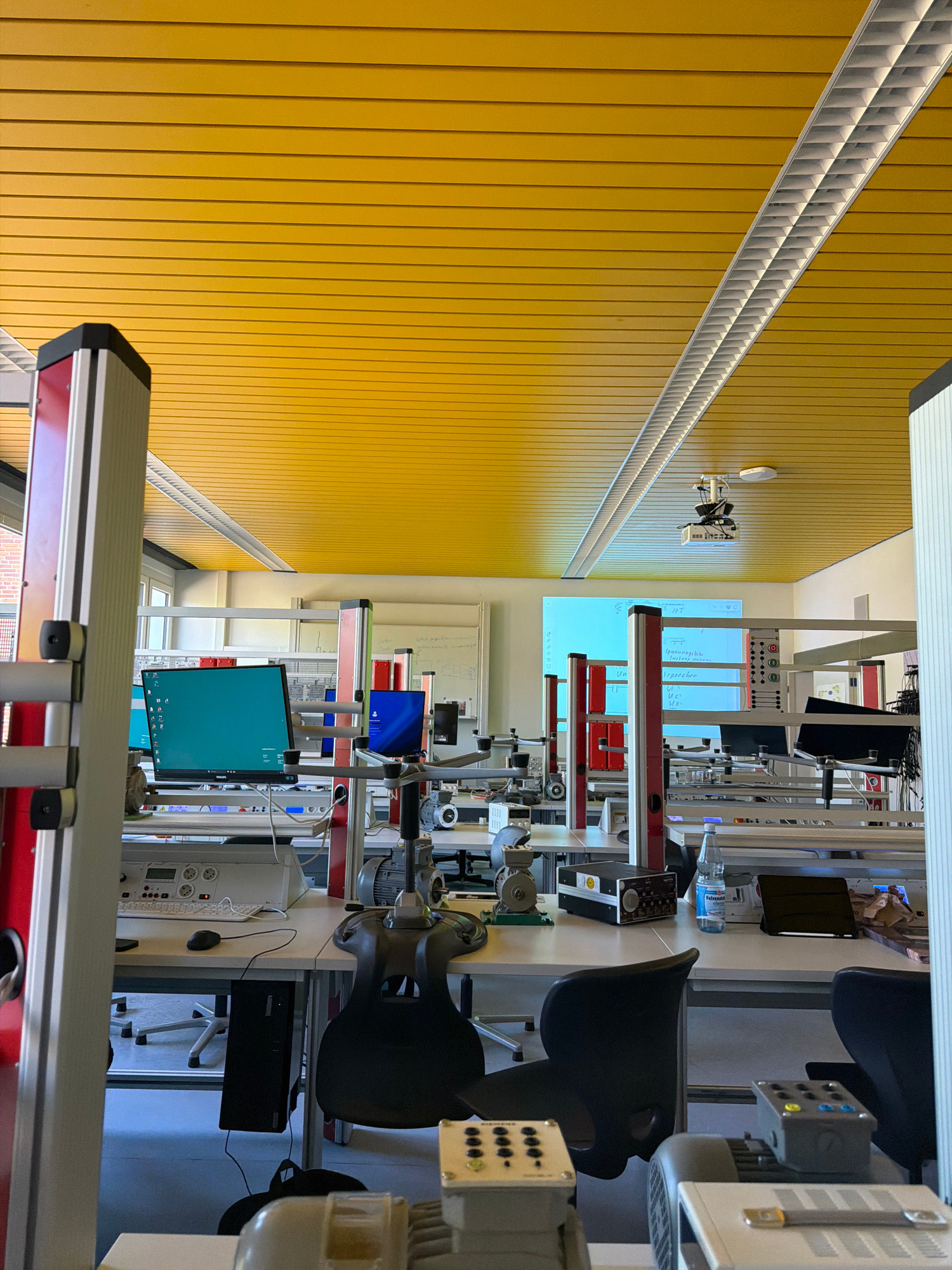 This image depicts a classroom or laboratory setting, likely within a vocational school or university in Warendorf, Germany, given the presence of technical equipment and educational materials. The room features a bright yellow, slatted ceiling with fluorescent lighting fixtures. Several workstations are visible, each equipped with computers, monitors displaying various interfaces, and scientific or technical apparatus, including motors and control panels. Desks are organized in rows, with some featuring chairs. A projector is mounted on the ceiling, pointing towards a projection screen displaying text and diagrams. The overall environment appears organized and functional, geared towards practical learning and experimentation. There are no individuals visible in the image.