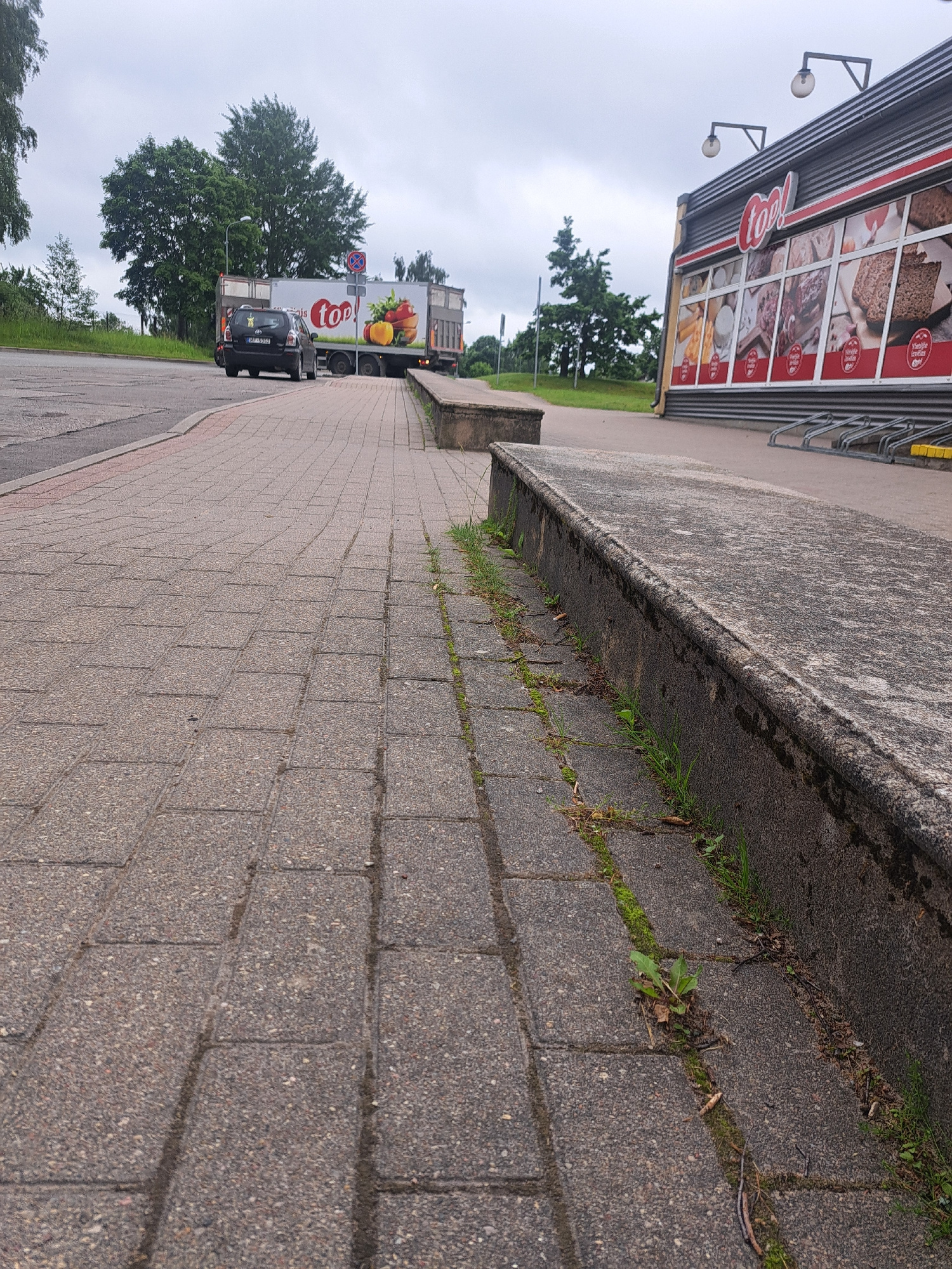 An exterior, ground-level, low-angle shot captures a sidewalk made of grey interlocking pavers in front of a "TOP!" grocery store in Cēsis, Latvia. The store's facade features a red trim and a large sign with the "TOP!" logo. Large windows display images of baked goods and food products. Adjacent to the store, a concrete barrier runs along the edge of the sidewalk, with some grass and weeds growing through cracks. In the background, a black car is parked next to a delivery truck, which is also branded with the "TOP!" logo and features an image of colorful peppers. The truck is parked on a paved area, possibly a parking lot, with a grassy embankment and trees visible behind it. The sky is overcast and a pale grey.