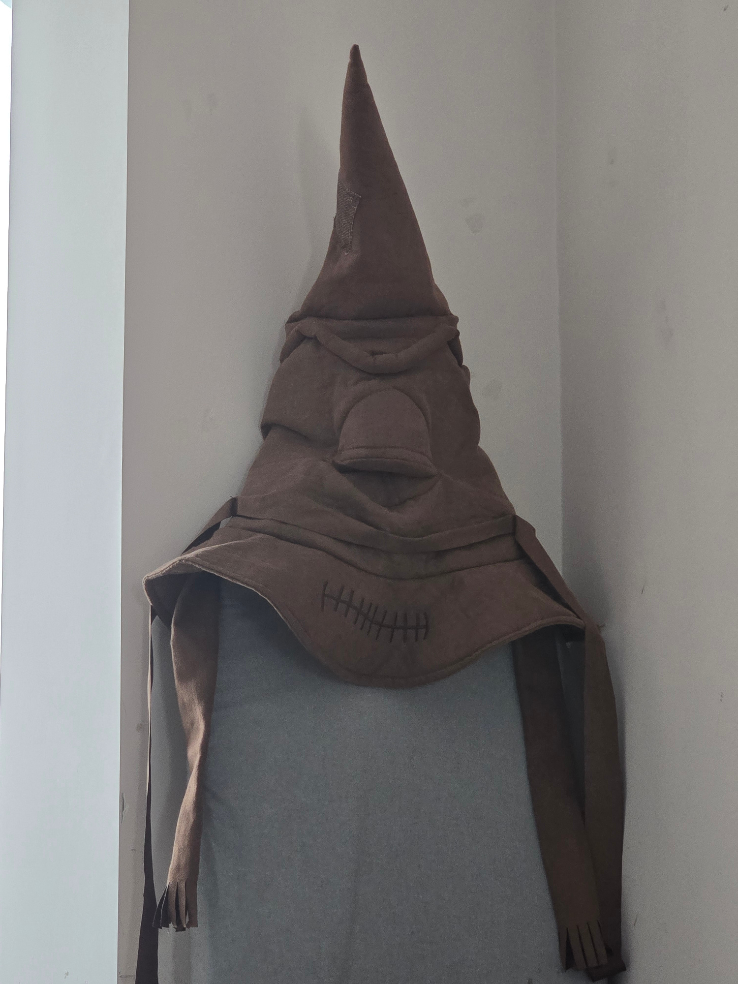 The image shows a brown, conical hat with ear-like flaps, resembling the Sorting Hat from Harry Potter. The hat is placed on what appears to be a gray surface, possibly a mannequin or a stand, in a corner formed by two walls. The hat has stitched details that suggest a face, with lines for a mouth. The overall scene is dimly lit. There is no indication of the location being in Petropavlivska Borshchahivka, Ukraine, or any other city.