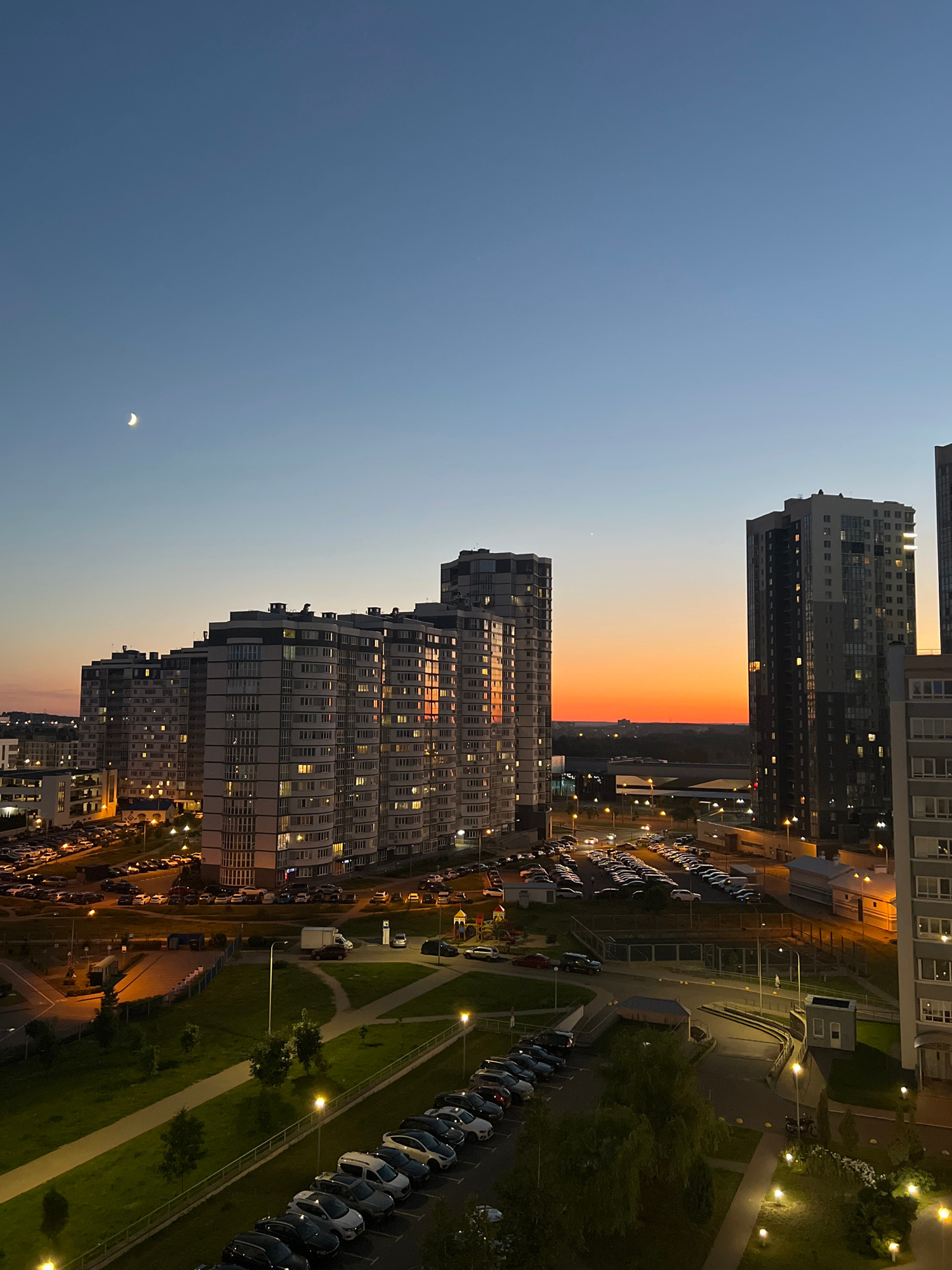 This vertical, eye-level, medium shot captures a cityscape at dusk, featuring modern high-rise apartment buildings and a vibrant sunset sky. The sky transitions from a deep blue at the top to a bright orange and yellow band just above the horizon, with a sliver of the moon visible in the upper left quadrant. A row of tall, multi-story residential buildings dominates the middle ground, their windows illuminated, casting a warm glow. In the foreground, there is a parking lot filled with numerous cars, and a section of a green park with lampposts. The scene suggests a residential district within the city of Minsk, Belarus, given the architectural style of the buildings. There are no discernible people or animals in the frame. The overall impression is one of urban tranquility at the close of day.