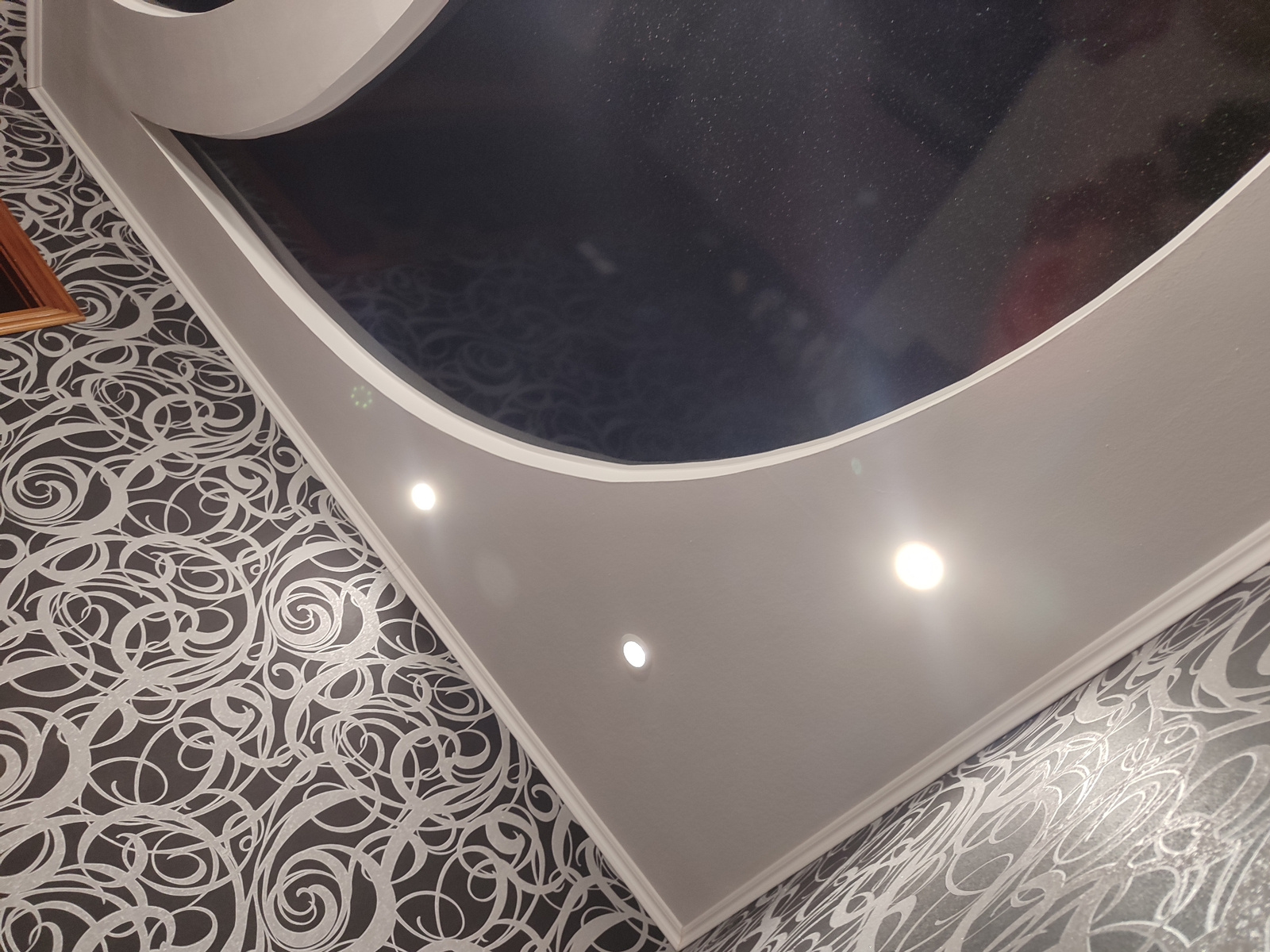 The image shows a ceiling with a modern design, featuring a dark, possibly reflective central area surrounded by a white, curved structure. Several recessed lights are visible on the white structure. The surrounding walls are covered in patterned wallpaper. The photo appears to be taken from a low angle, looking upwards. There are no people or specific actions depicted in the image. The scene is indoors and likely a residential or commercial space. No specific location in Mykolaiv, Ukraine, can be determined from the image alone.